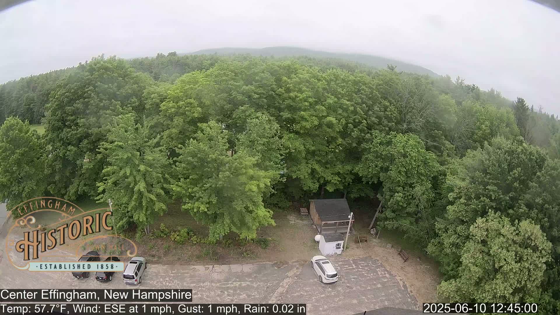 A high-angle, long shot shows a light rain falling on a parking lot, two cars, and a small shed surrounded by a lush green forest under an overcast sky.