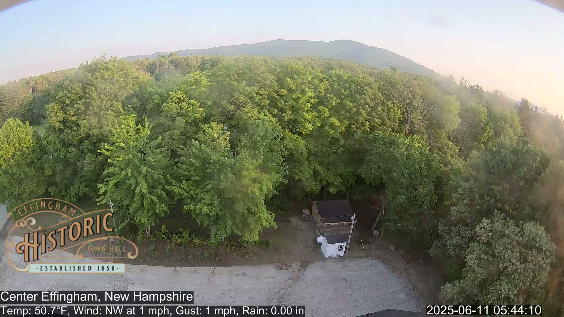 An aerial view shows a small shed and lush green trees under a mostly clear sky, with a mountain range visible in the distance.