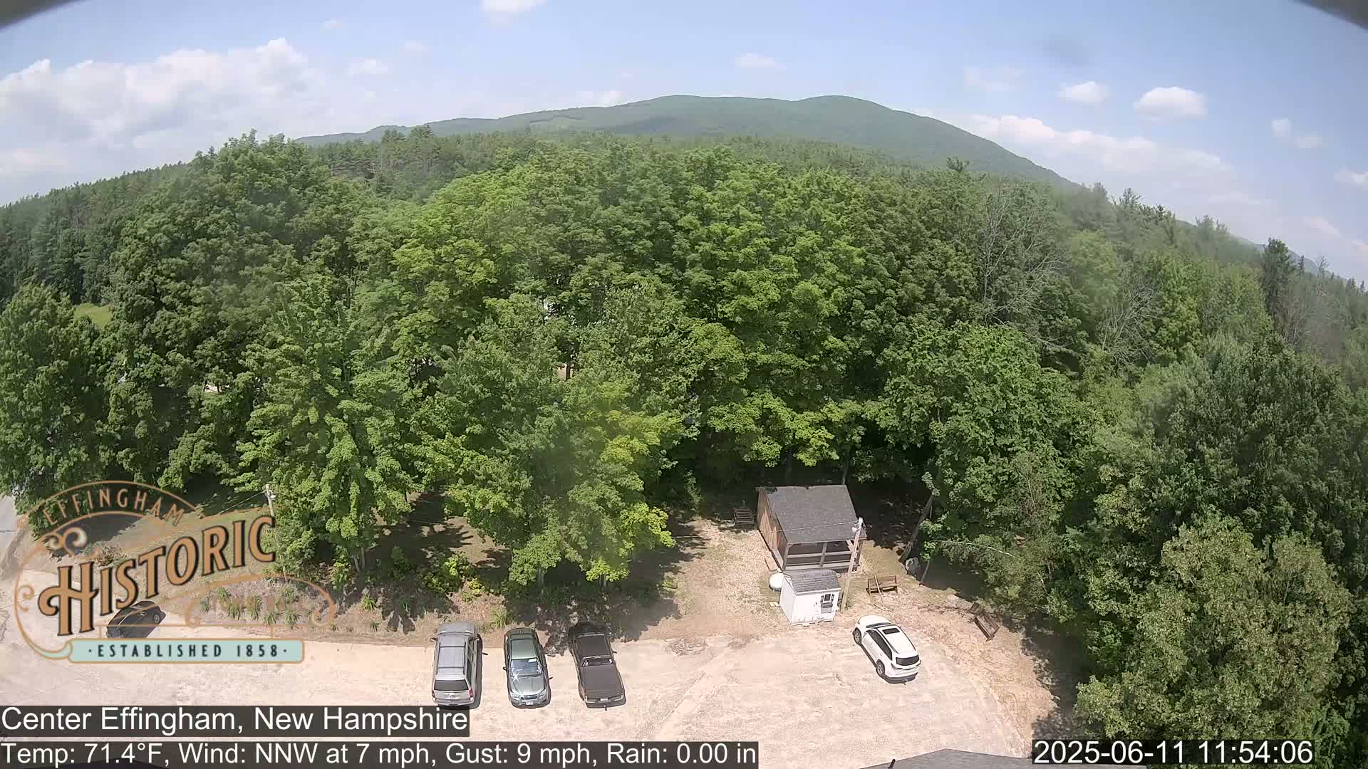 An aerial view shows a parking lot with several cars next to a small building surrounded by lush green deciduous trees and a distant mountain range under a mostly clear sky.