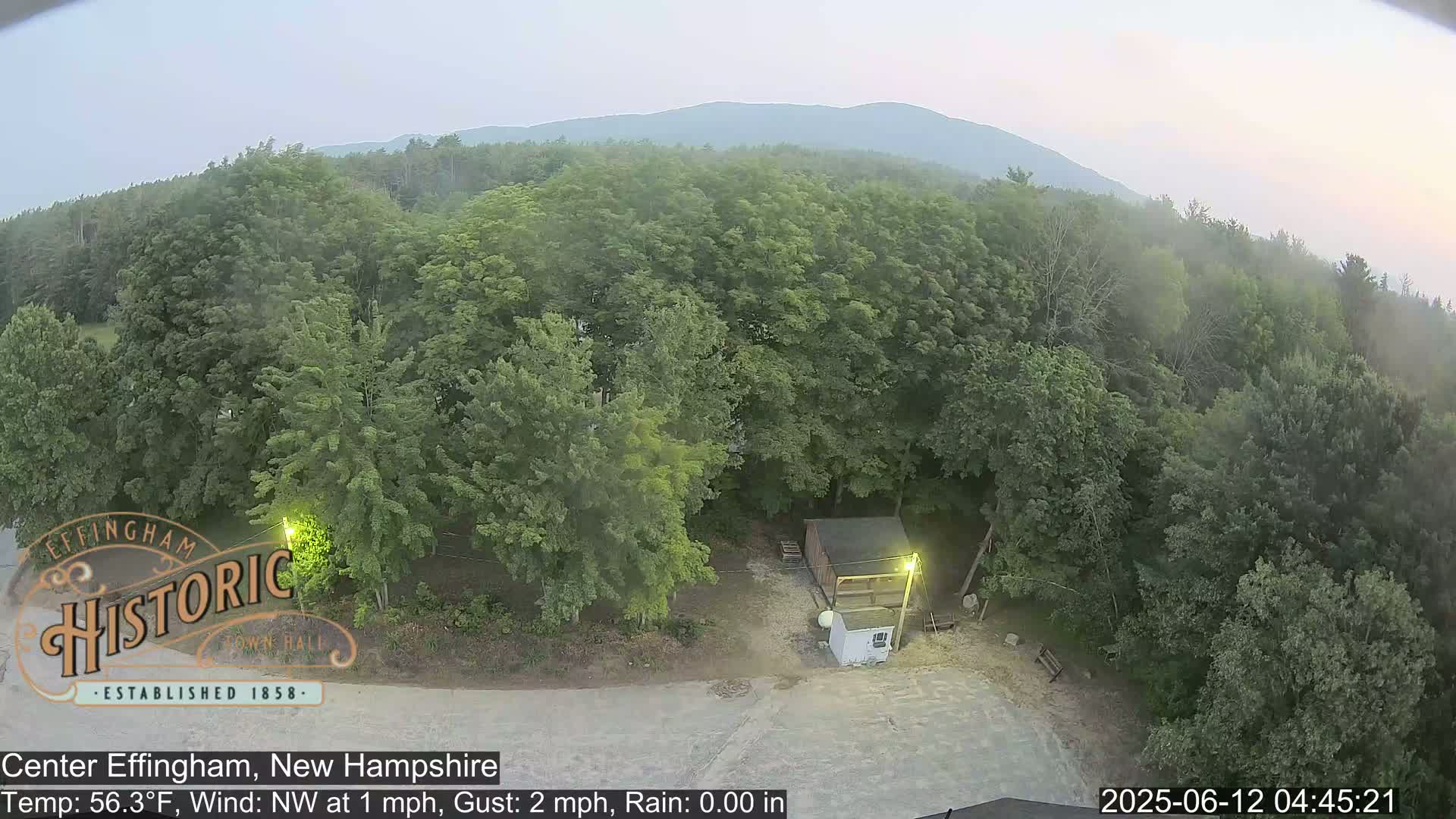 An elevated view shows a small building in a clearing surrounded by lush green trees under a hazy sky.