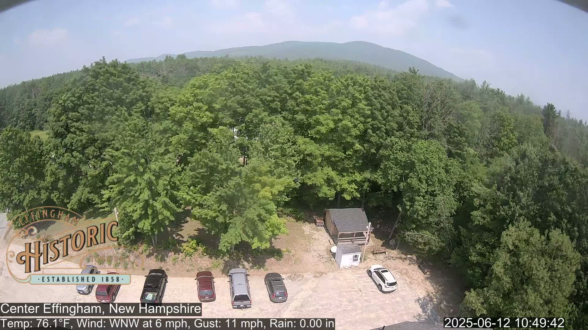 An aerial view shows several cars parked in a lot next to a small building surrounded by lush green trees and a distant mountain range under a partly sunny sky.