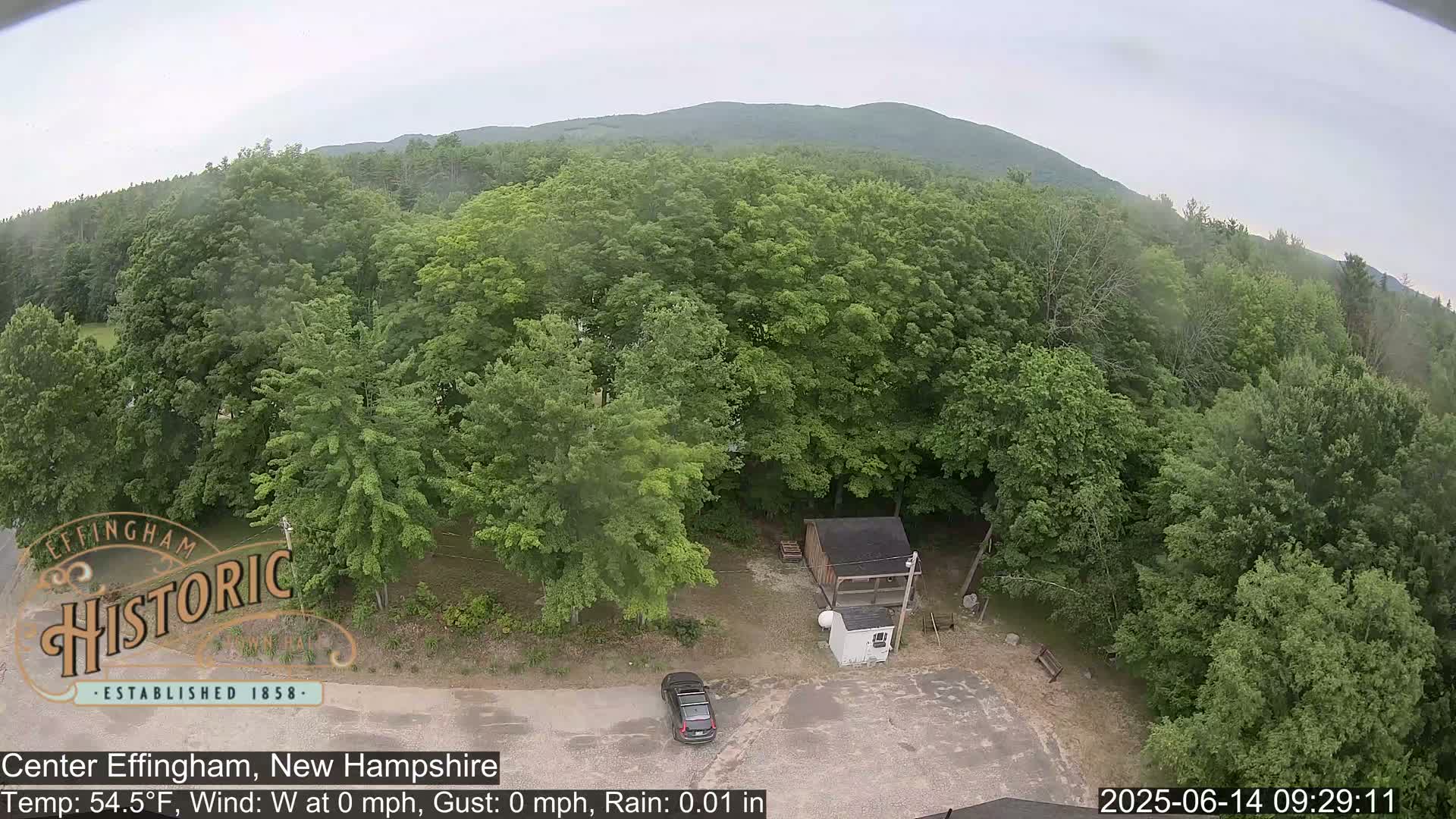 An aerial view shows a small parking lot with a car next to a small building surrounded by lush green trees and a distant mountain range under slightly overcast skies with light rain.