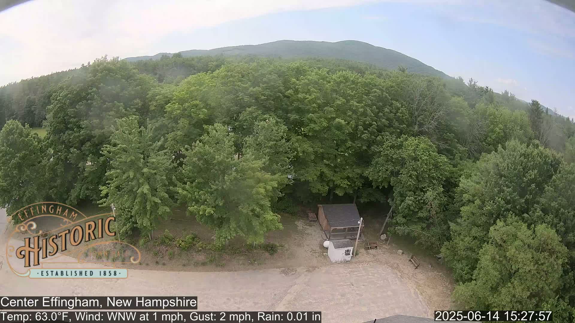 An aerial view shows a small parking lot with a car next to a small building surrounded by lush green trees and a distant mountain range under slightly overcast skies with light rain.