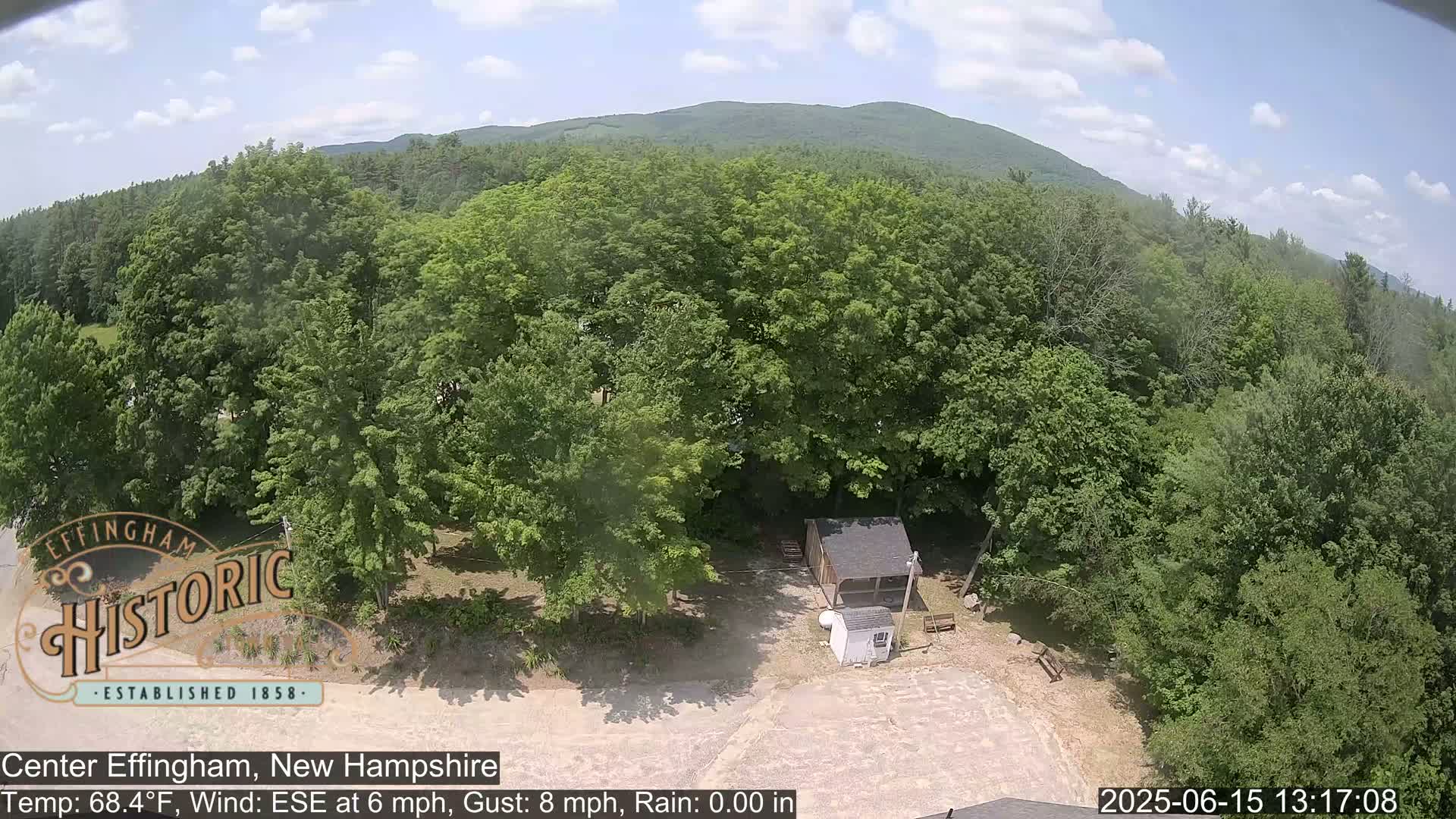 An aerial view shows a mostly sunny day over a wooded area with a small shed and camper, a mountain in the background.