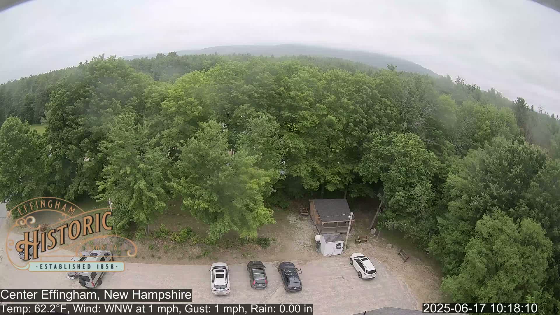 A high-angle, slightly overcast view shows a parking lot with several cars next to a small building surrounded by lush green trees.