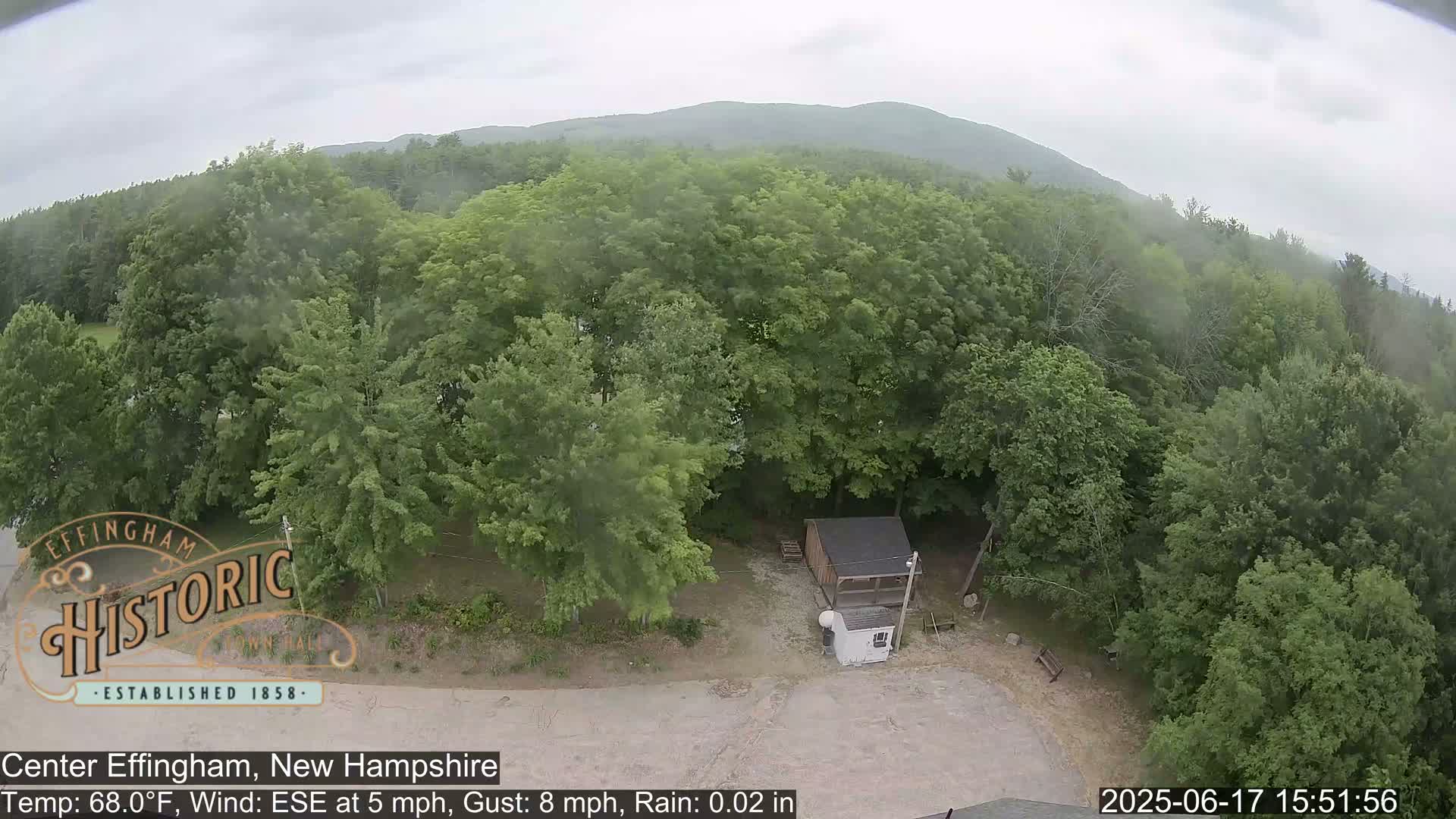 An aerial view shows a small shed and a parking area surrounded by lush green trees under a lightly overcast sky with light rain.