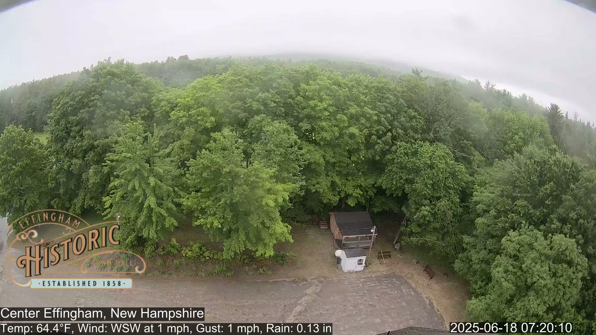 An aerial view shows a small building and a paved area surrounded by lush green trees in light rain.