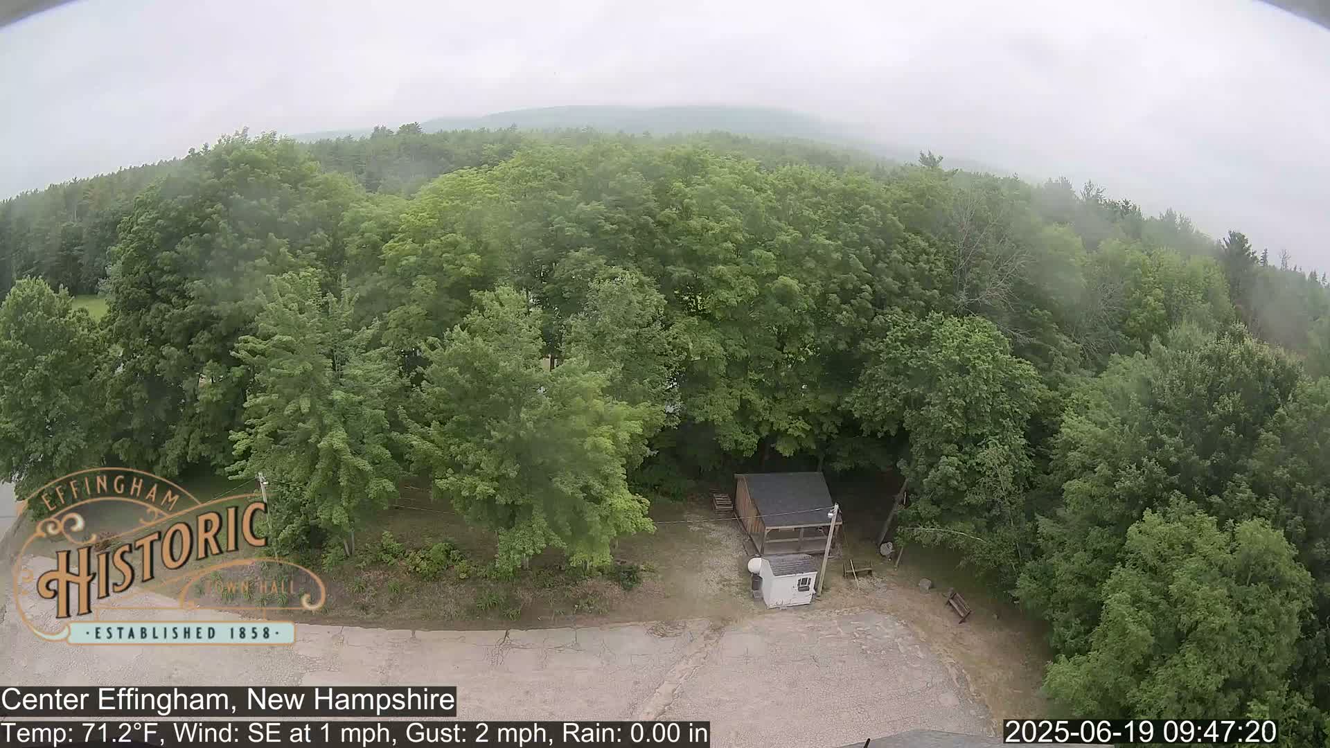 An aerial view shows a small structure nestled amongst lush green trees on a slightly overcast day with light rain.