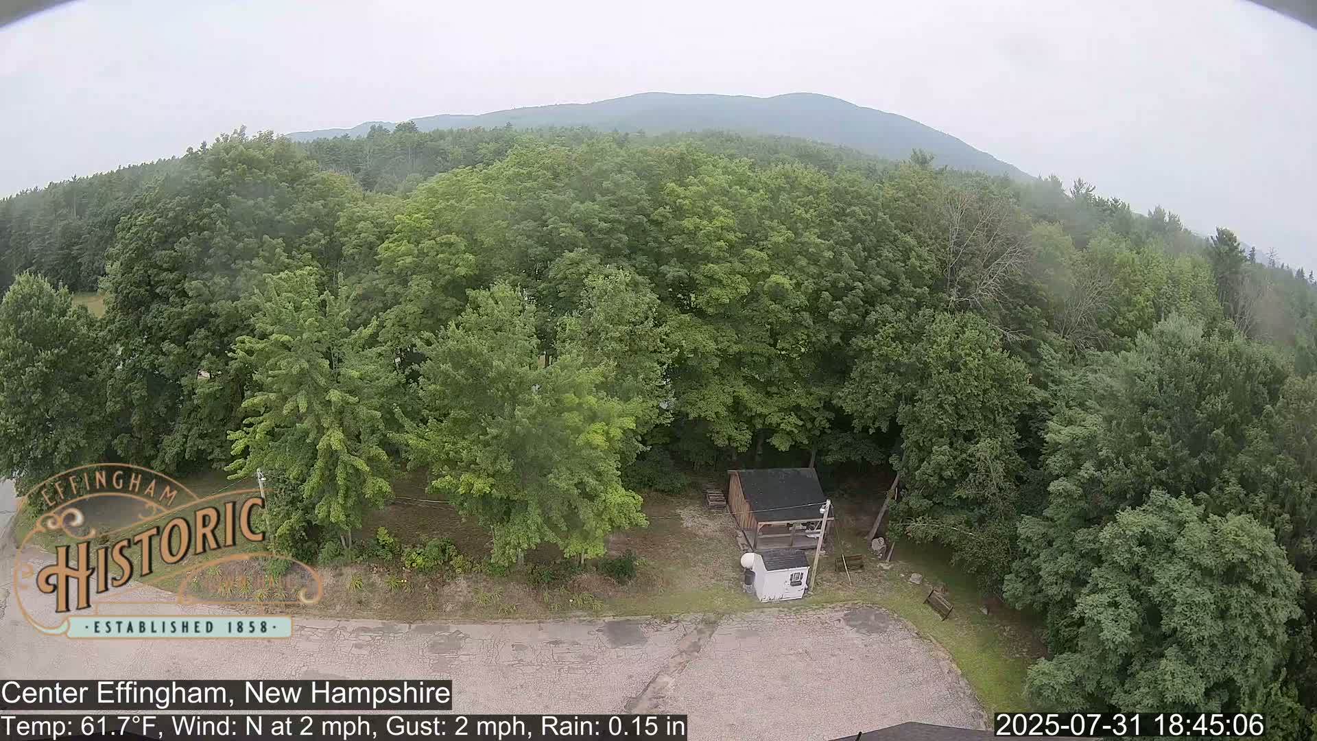 An aerial view shows a small building and surrounding lush green trees under rainy conditions, with a mountain in the distance.