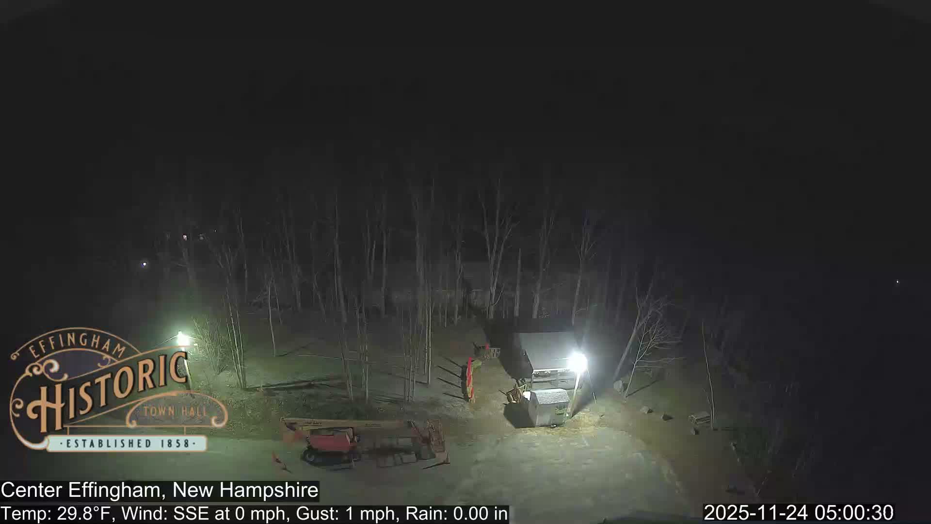 A dark outdoor scene at night features a sparse forest of bare trees, a red boom lift, and a brightly illuminated shed, under clear and calm weather conditions.