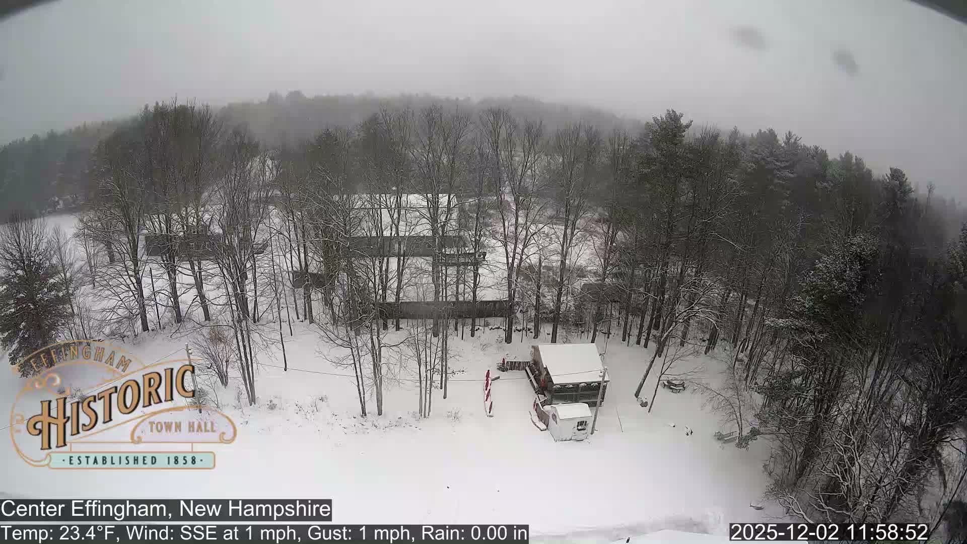 A wide, high-angle view captures a snow-covered winter landscape featuring numerous bare trees, scattered buildings, and distant hazy hills under a uniformly grey and overcast sky.