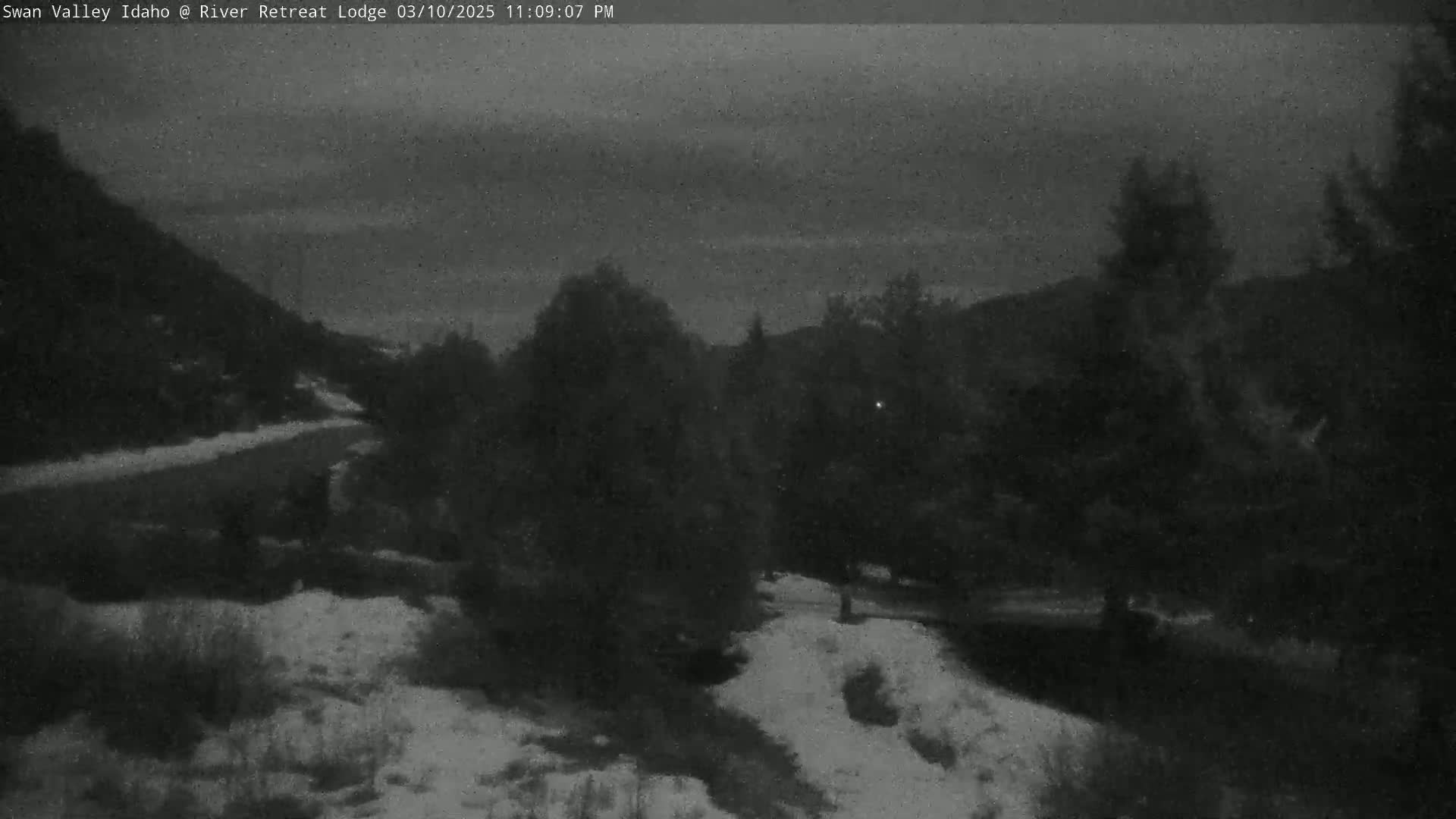 A nighttime view of a snow-dusted landscape, featuring a winding road through dark trees and hills under a cloudy sky.