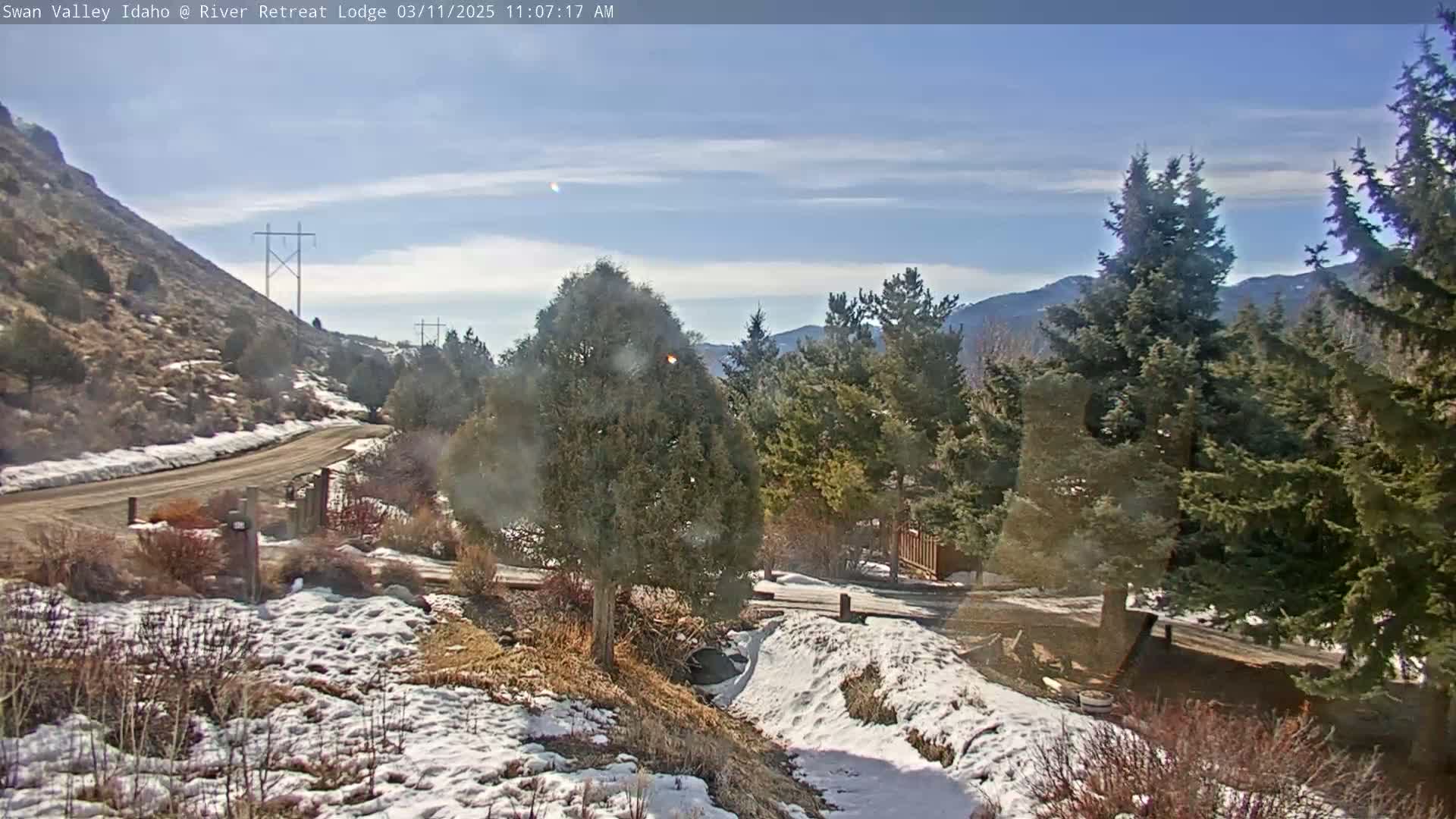 A partly sunny day with snow on the ground shows a dirt road winding through a landscape of evergreens, shrubs, and a snow-dusted hillside.
