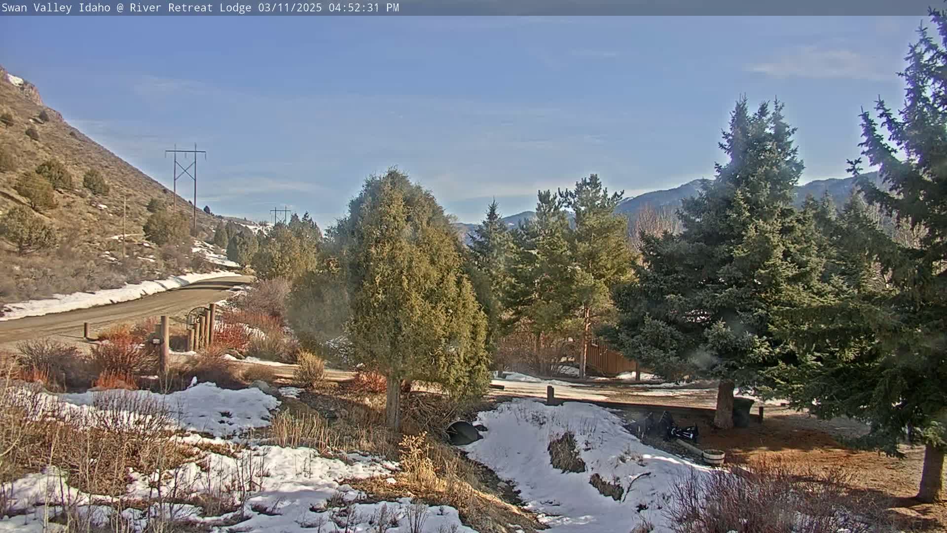 A partly sunny day reveals a snow-dusted landscape with evergreen trees, a dirt road, and a hill in the background.