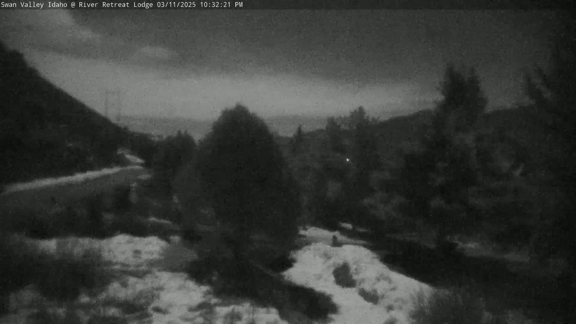 A nighttime, snow-dusted landscape shows dark trees and hills under a cloudy sky.