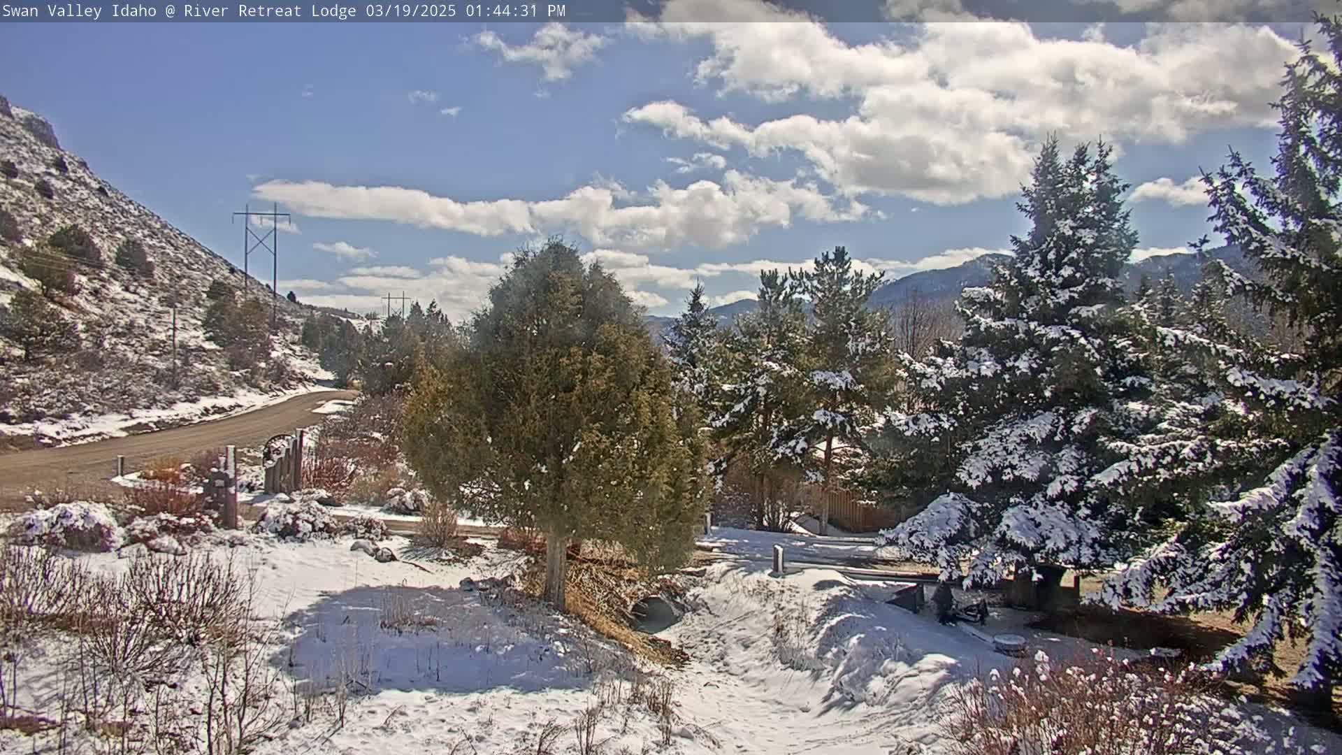 A snow-dusted landscape features a road winding through a valley, evergreen trees partially covered in snow, and a partly cloudy sky.