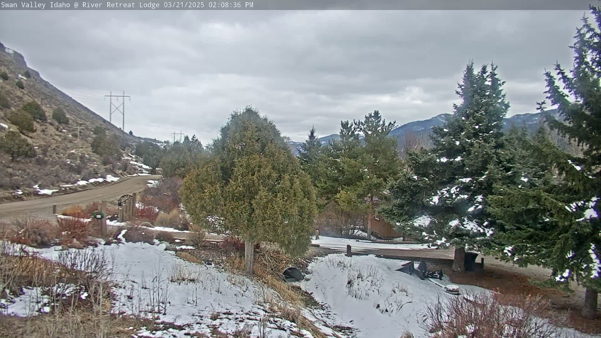 A snow-dusted landscape shows a dirt road curving past evergreen trees and a small building, with a hill rising in the background under a cloudy sky.