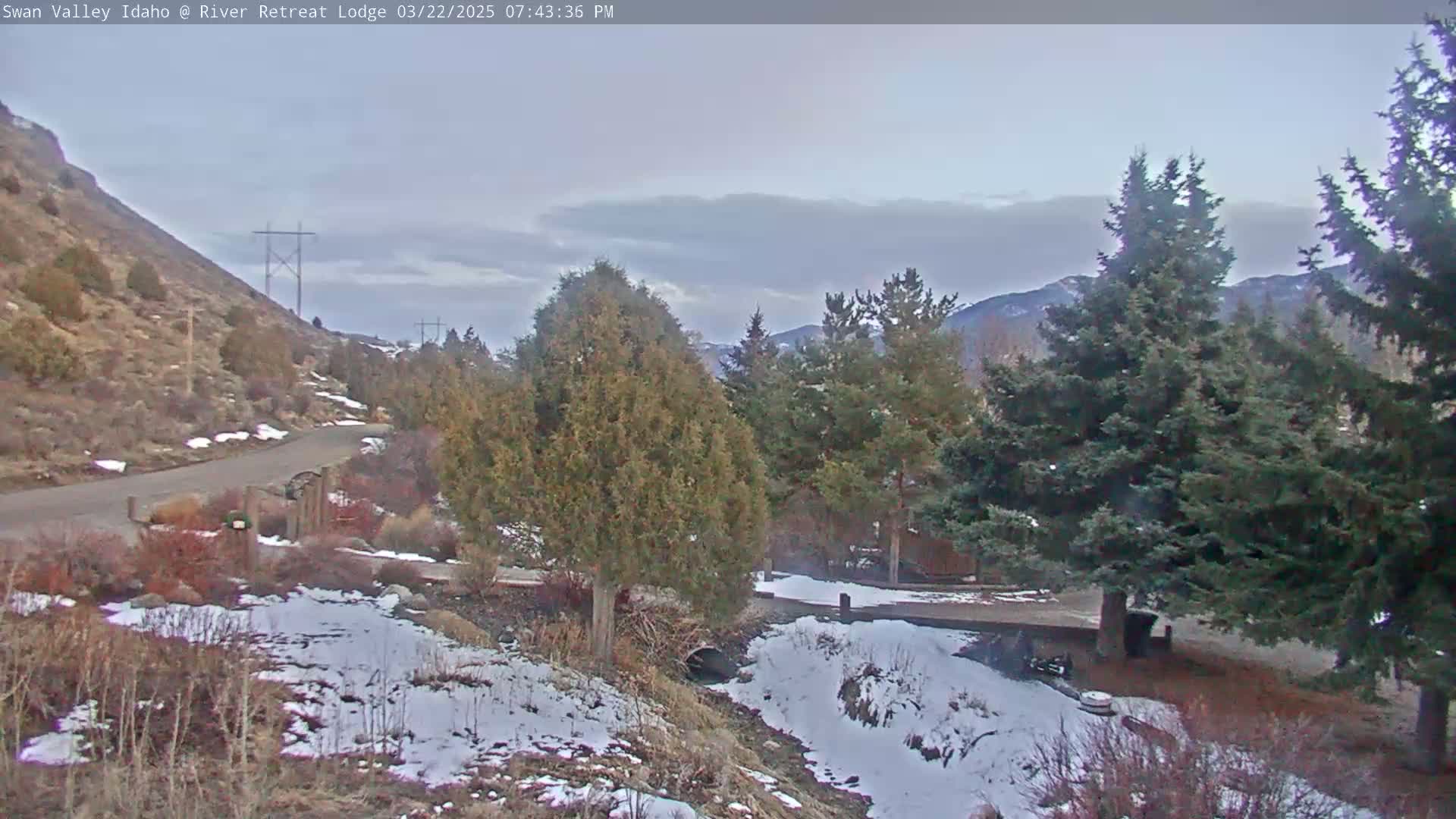 A paved road winds through a snow-dusted landscape of brown hills, evergreen trees, and a small stream, under a mostly cloudy sky.