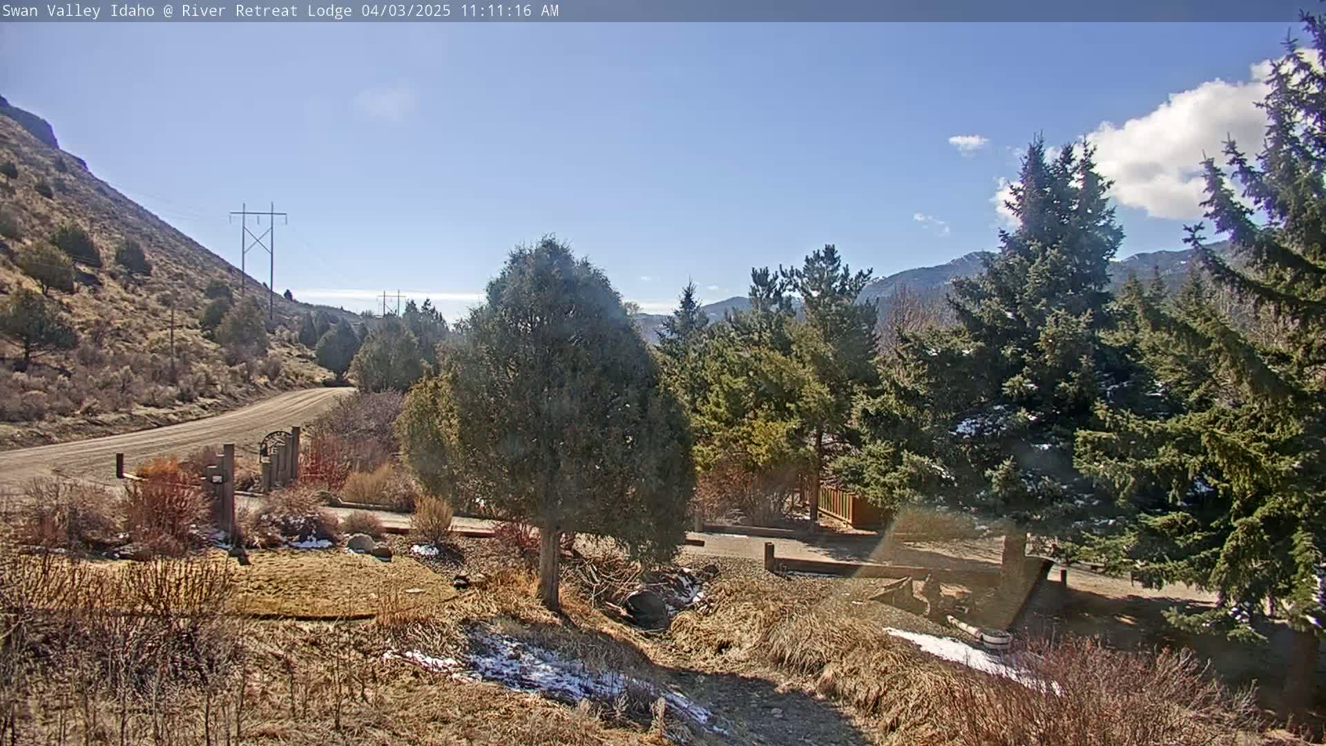 A sunny day reveals a dirt road leading to a gated entrance, nestled amongst evergreen trees and a brown, hilly landscape with patches of snow.
