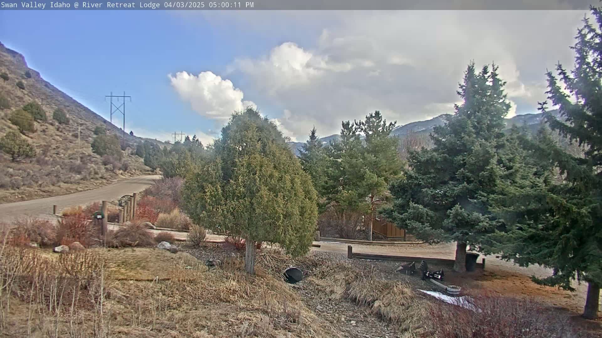 A partly cloudy day reveals a view of a gravel road leading to a lodge nestled among evergreen trees and a rocky hillside.