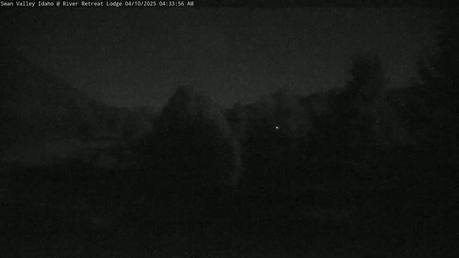 A dark, nighttime outdoor scene shows a dimly lit landscape with indistinct shapes suggesting trees or buildings under a mostly clear sky.