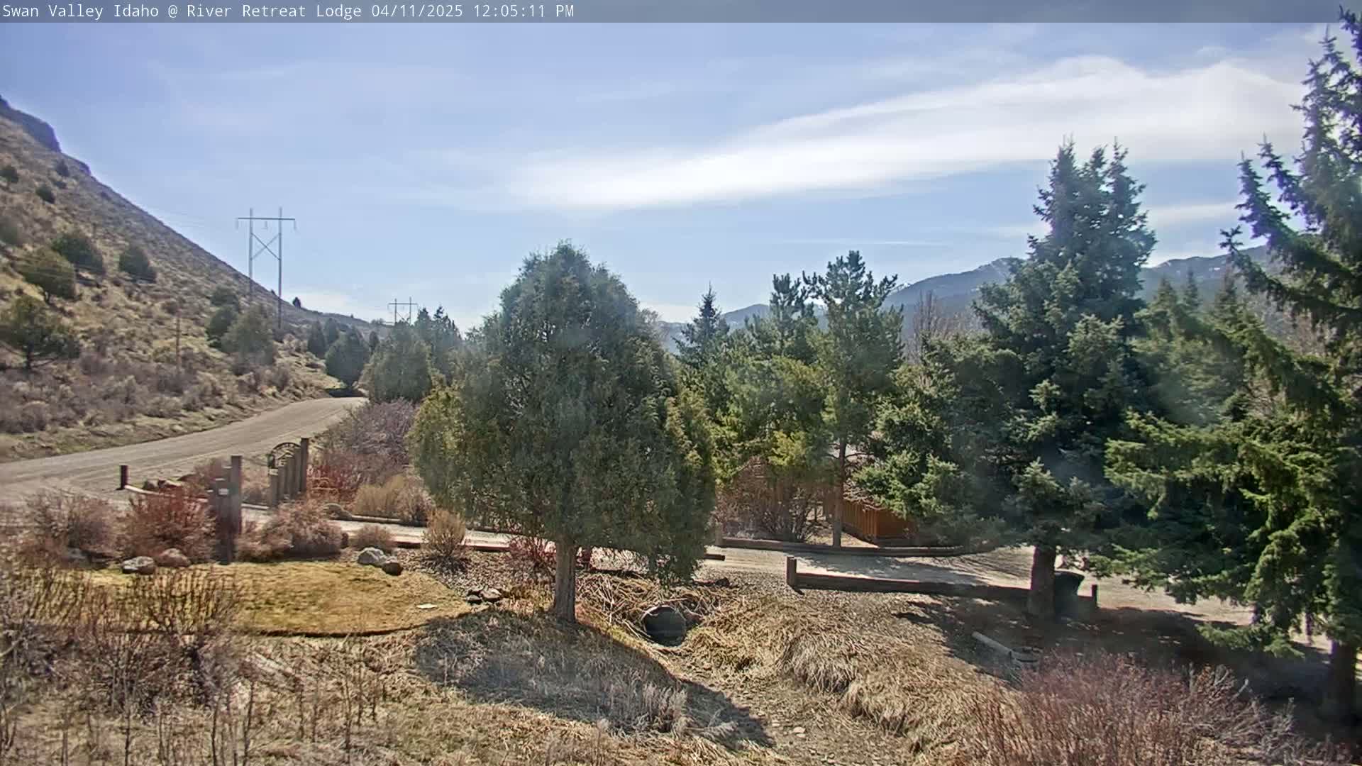 A gravel road leads to a gate, past landscaping and evergreen trees, with a partly cloudy sky and mountains in the background.