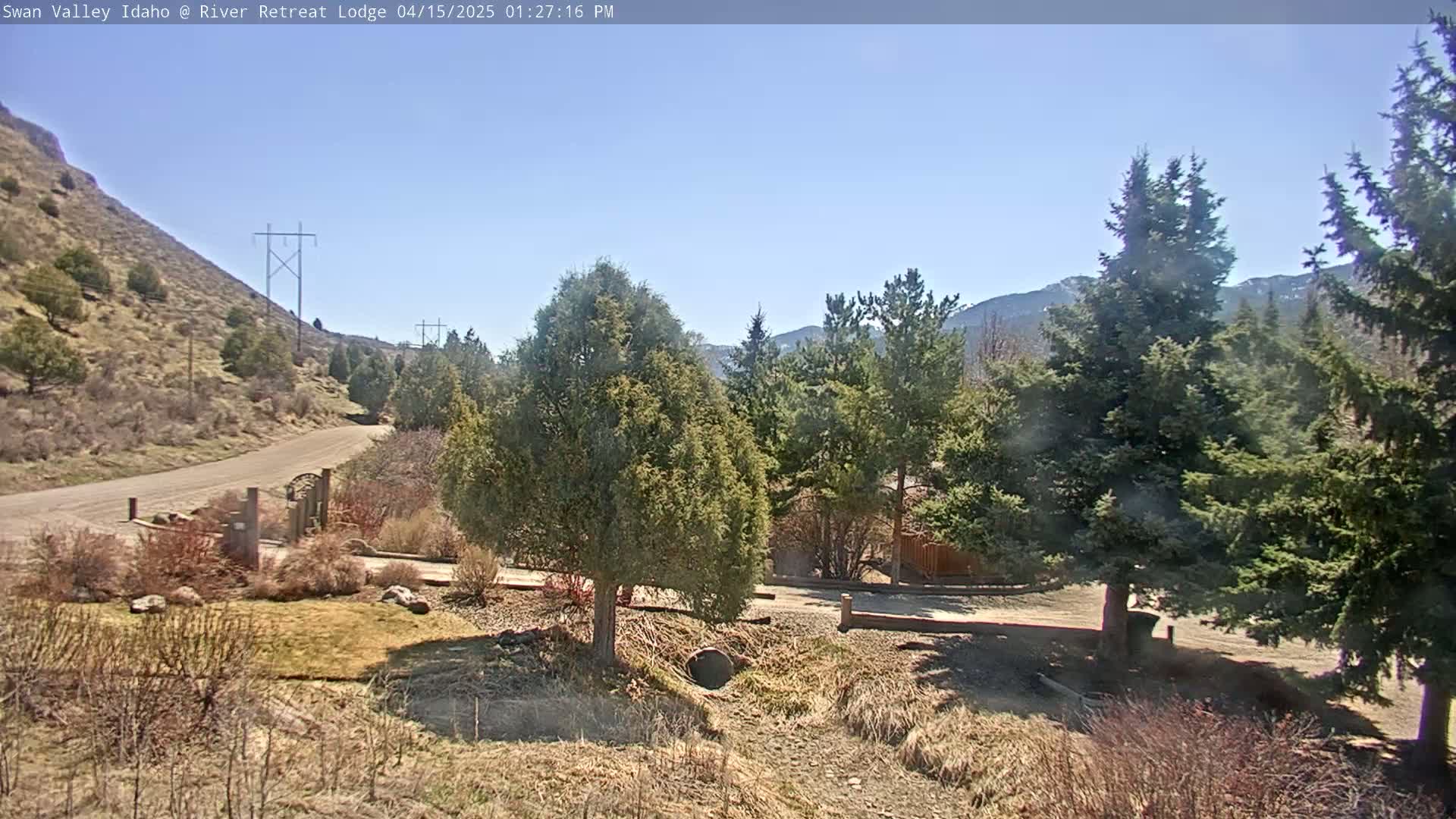 A gravel road winds through a mountainous landscape under a clear, sunny sky, passing evergreen trees and a small structure.