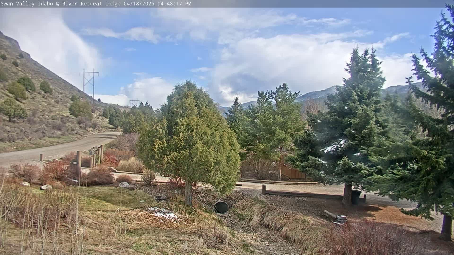 A partly cloudy day reveals a gravel road winding through a landscape of evergreen trees, hillsides, and a small drainage ditch, with a few patches of snow still visible.