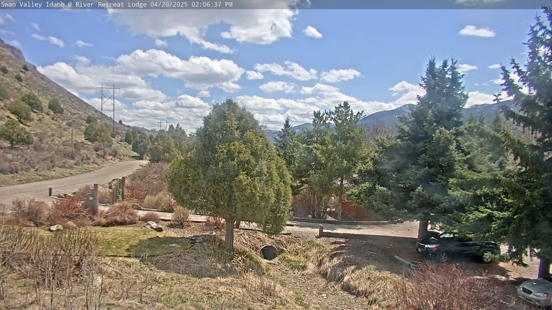 A partly sunny day reveals a winding paved road leading to a residential area nestled among evergreen trees and a hillside, with a dark-colored SUV parked nearby.