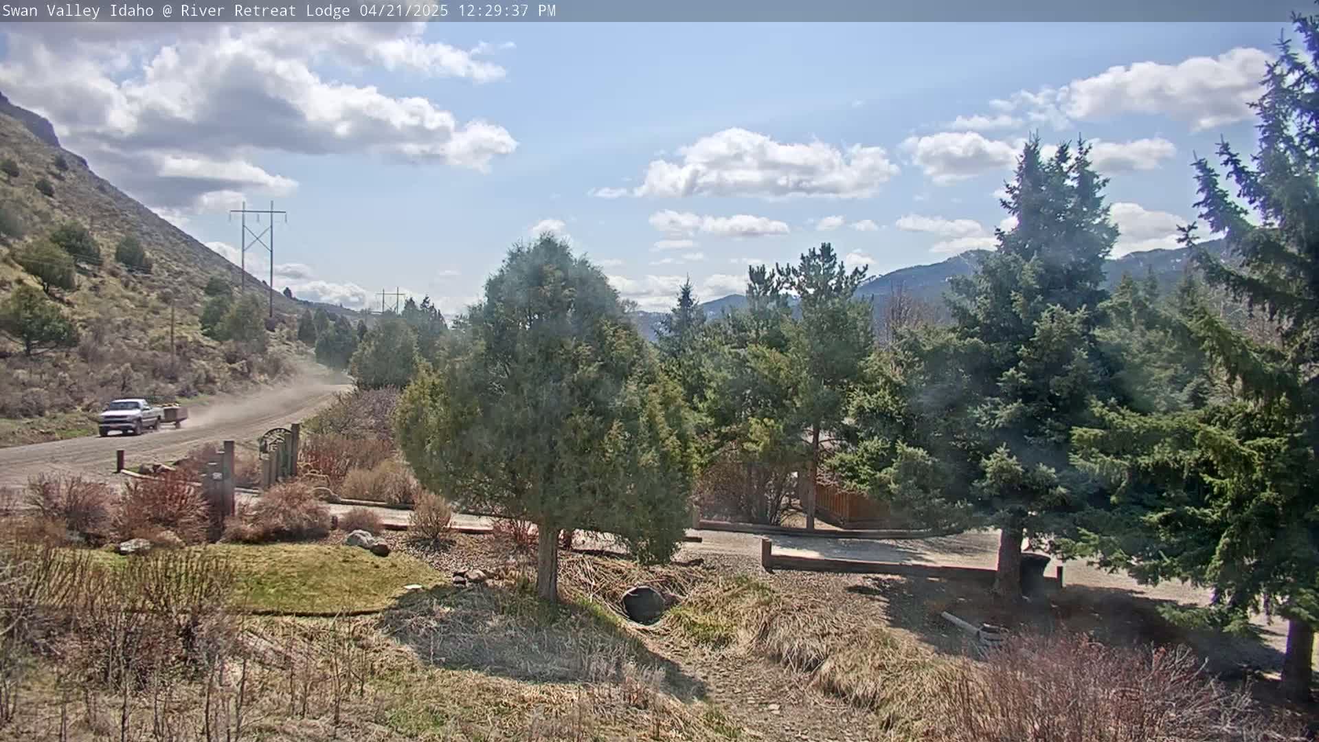 A partly cloudy day reveals a pickup truck driving down a dirt road past evergreen trees and a brown hillside, with mountains in the background.