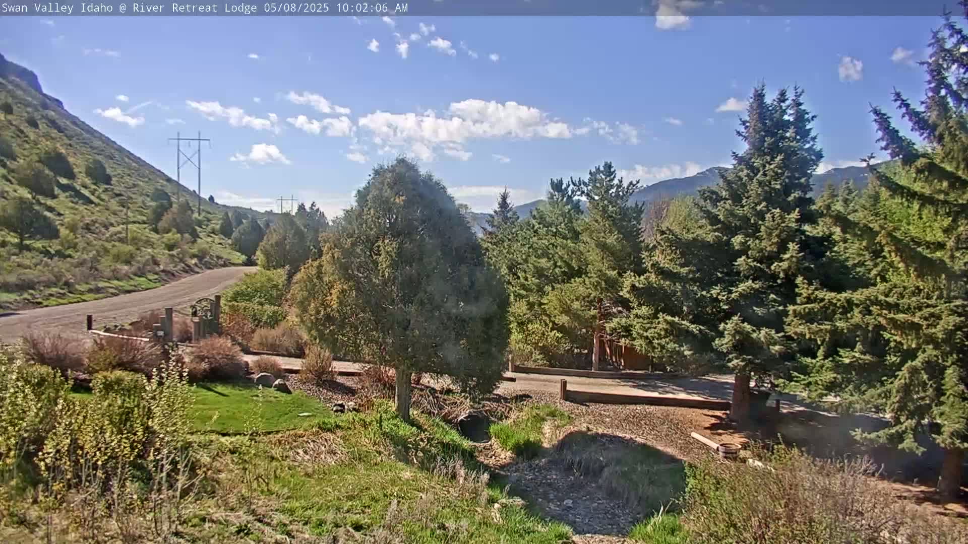 A gravel driveway leads to a lodge nestled among evergreen trees and a grassy hillside under a partly cloudy blue sky.
