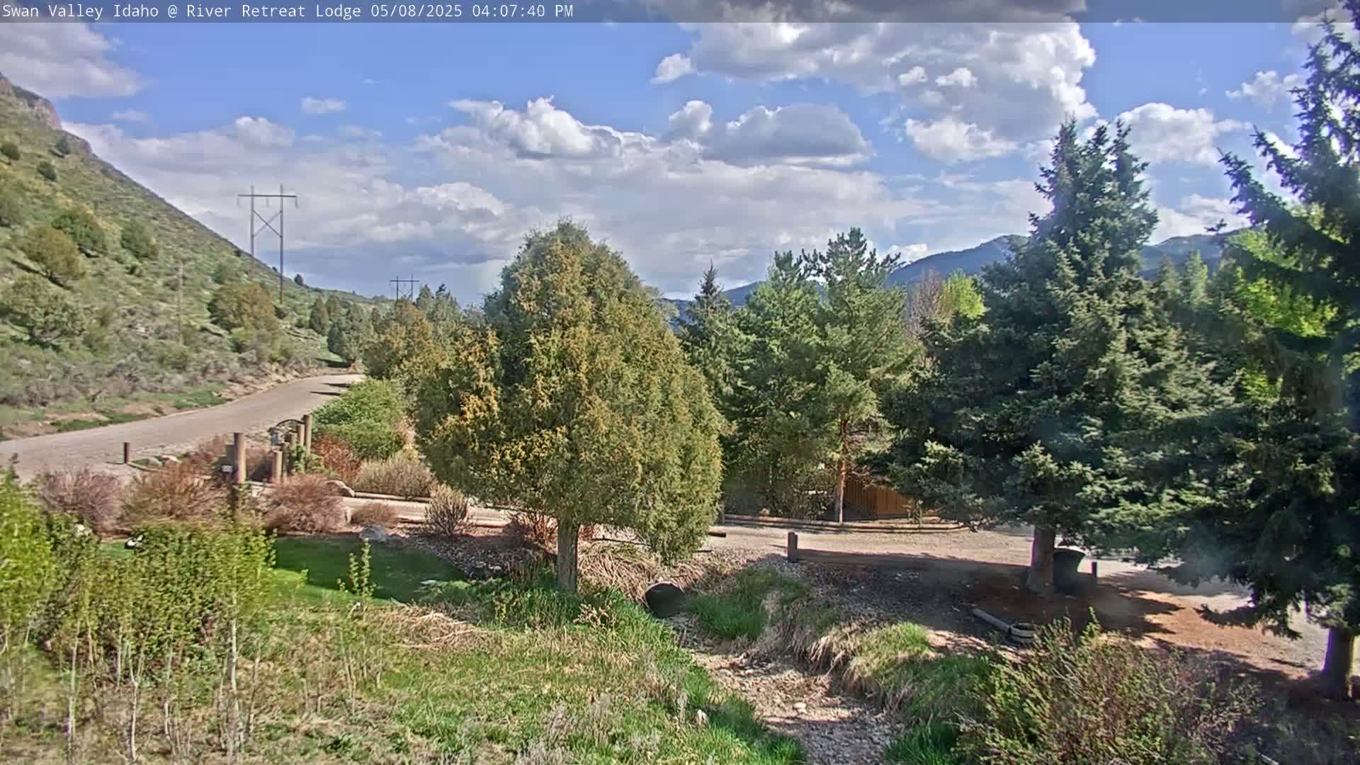 A partly sunny day shows a paved road winding through a landscape of evergreens and scrubby vegetation, with mountains in the background.