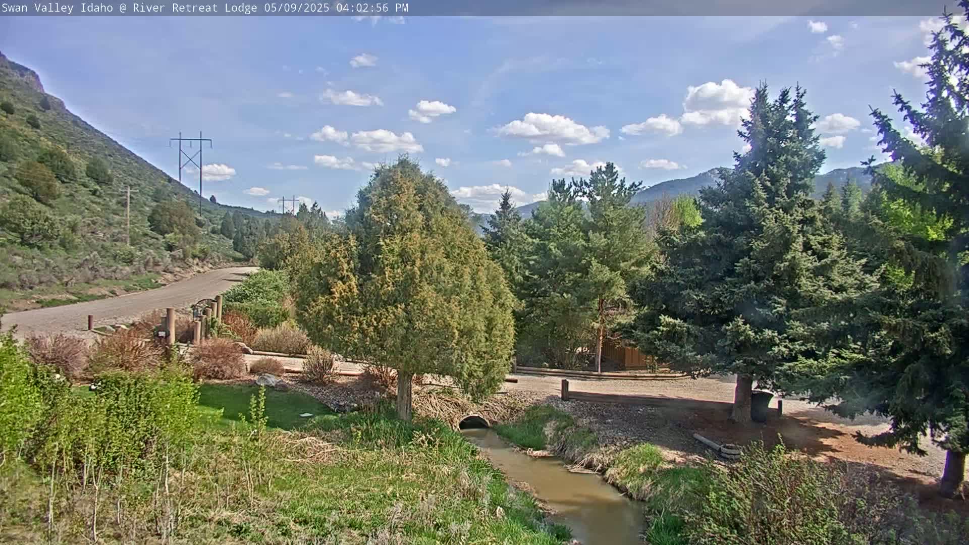 A paved road winds through a mountainous landscape under a partly sunny sky, with a small stream flowing beside it, lush greenery, and tall evergreen trees.