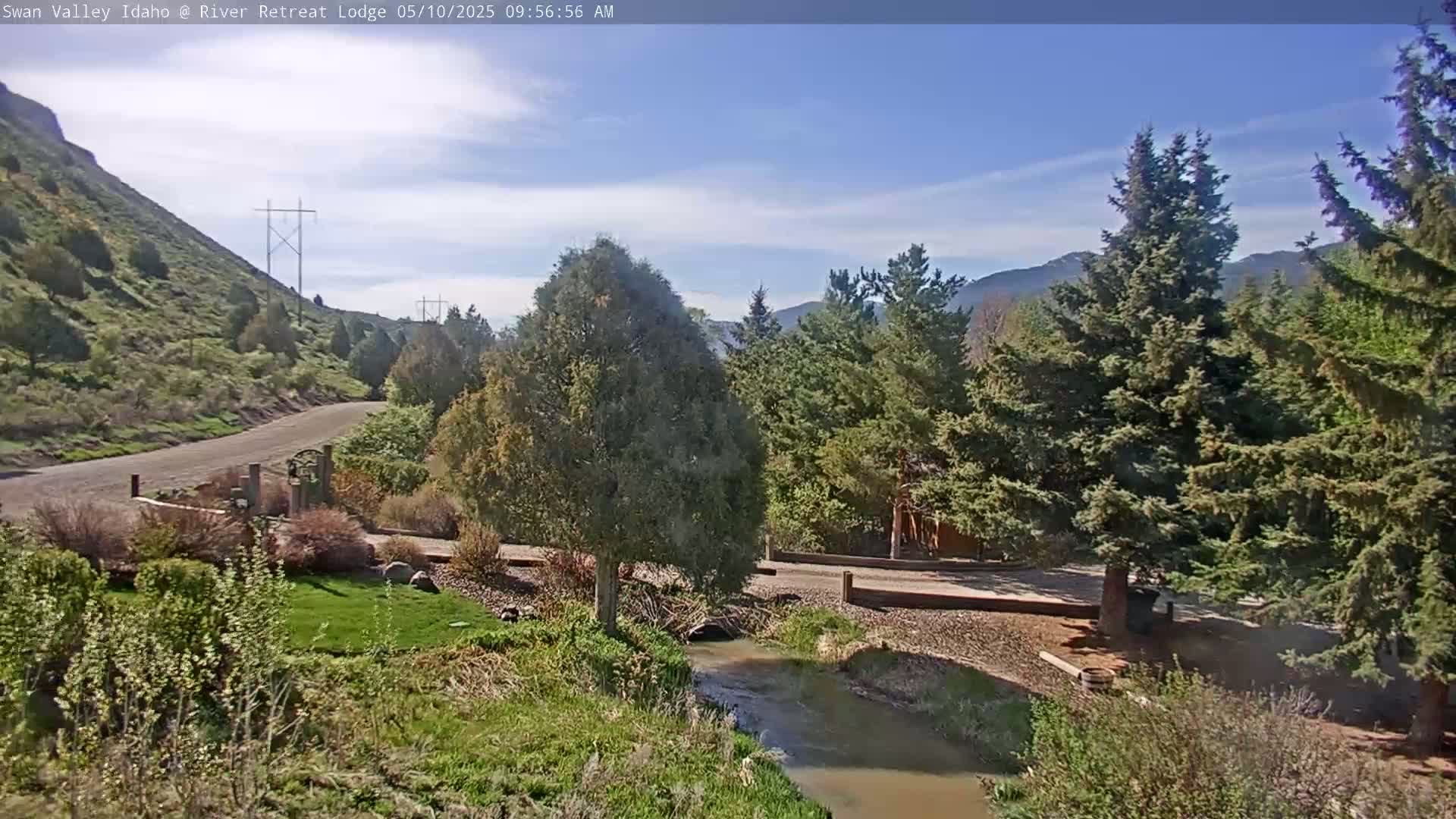 A gravel road winds through a green valley alongside a small stream, under a mostly sunny sky, with tall evergreen trees and a brown hill in the background.