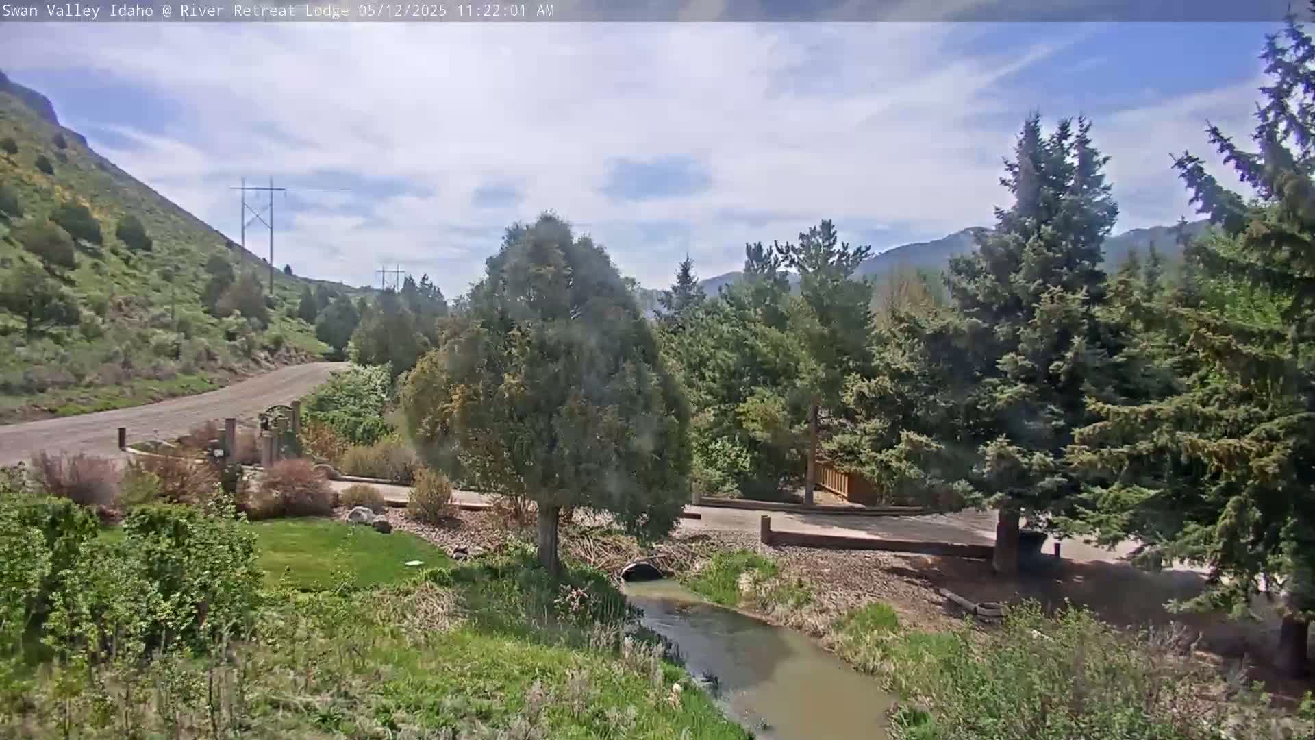 A paved road winds through a landscape of green hills, evergreen trees, and a small creek under a partly cloudy sky.