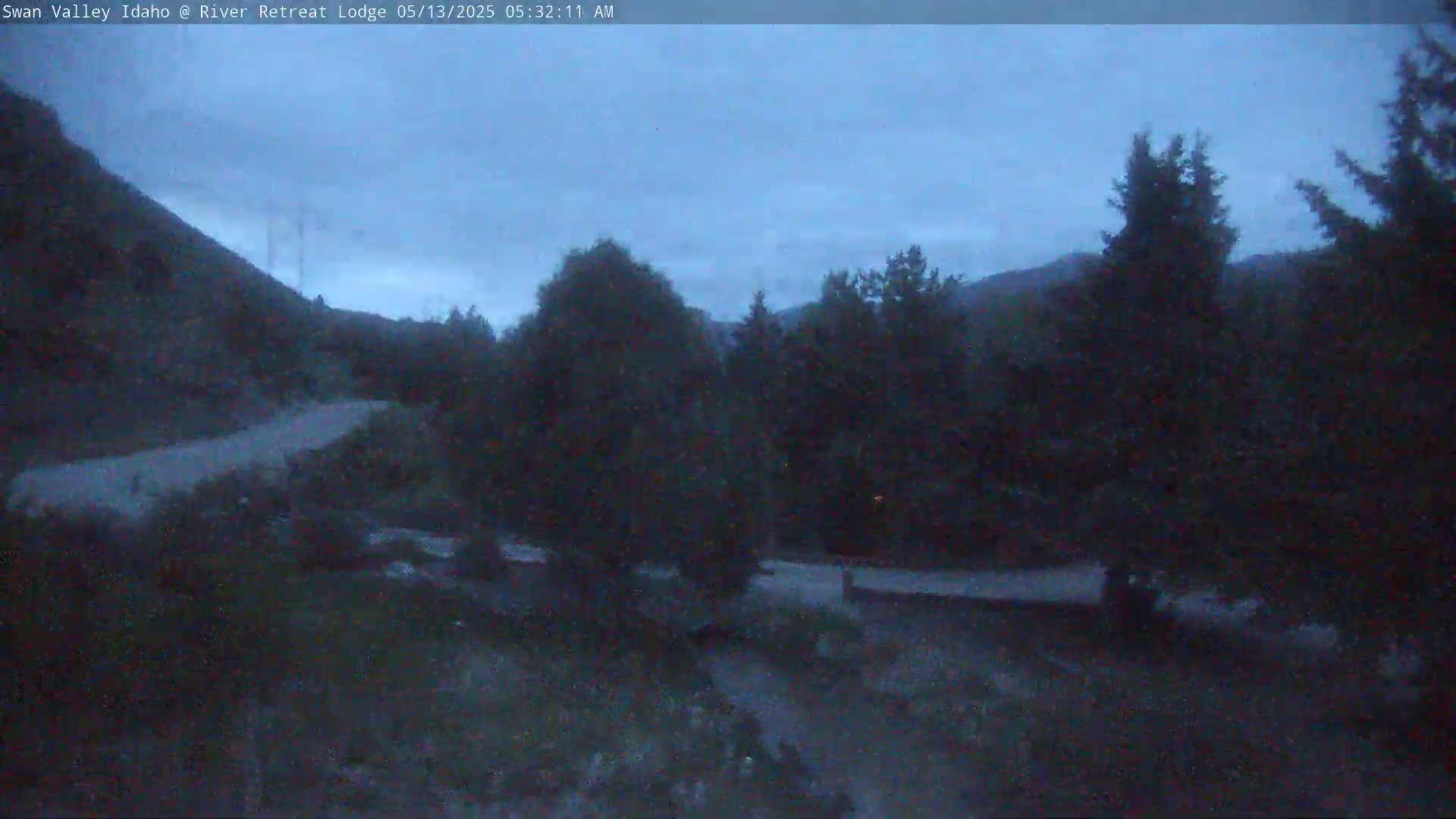 A dark, overcast morning scene shows a paved road winding through a mountainous area with dark green trees and shrubs.