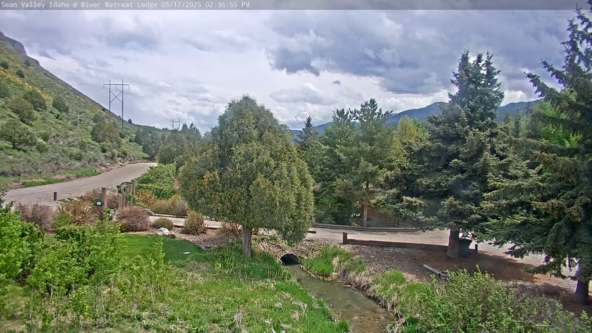 A paved road winds through a mountainous landscape under a partly cloudy sky, alongside a small stream flowing past lush greenery and evergreens.