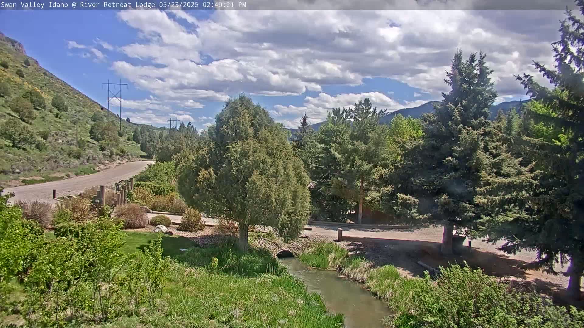 A partly cloudy sky overlooks a tranquil stream flowing through a green landscape with trees, a road, and a hill in the background.