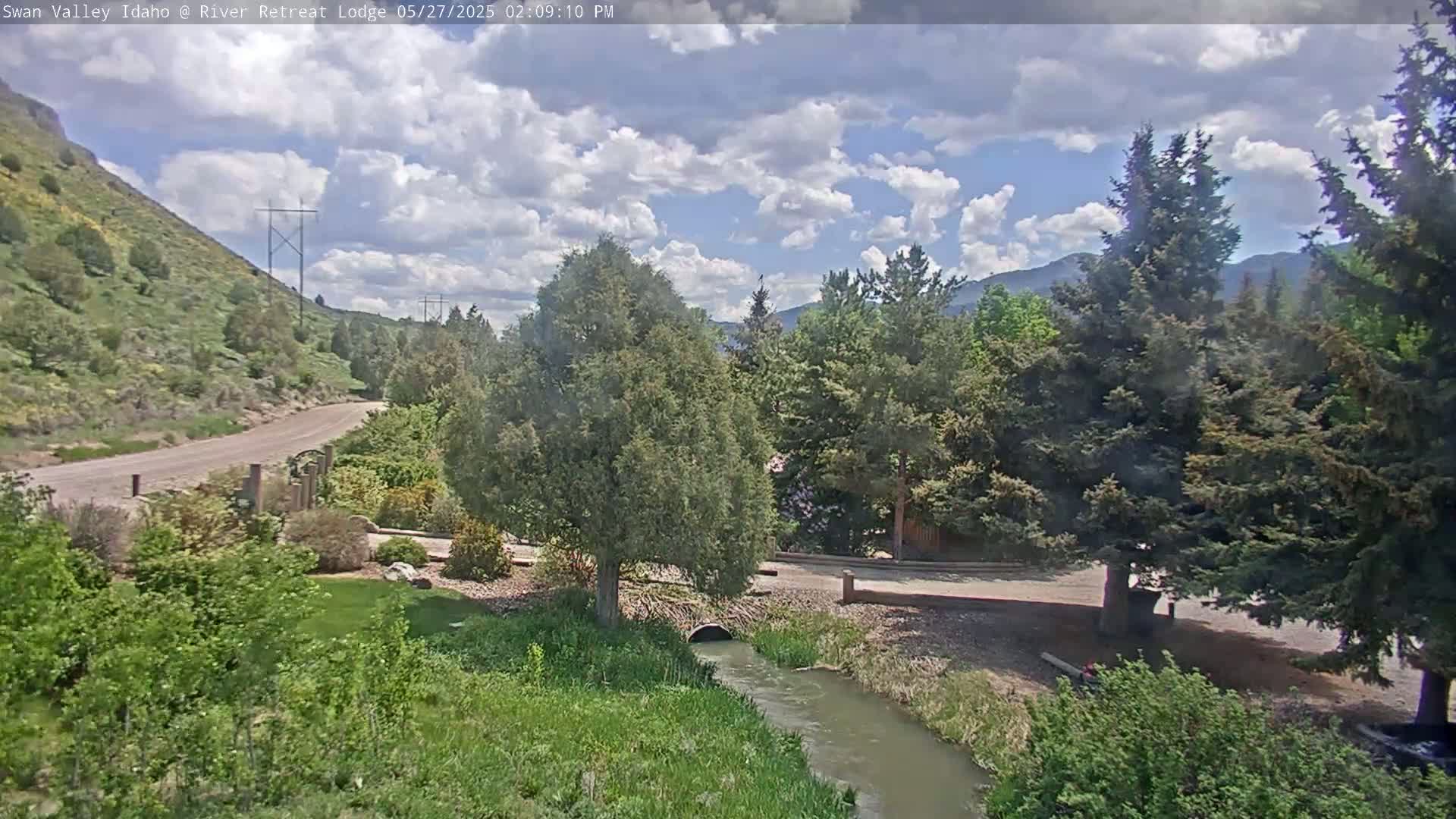 A small stream flows through a grassy area next to a road and lush green vegetation, under a partly cloudy sky.