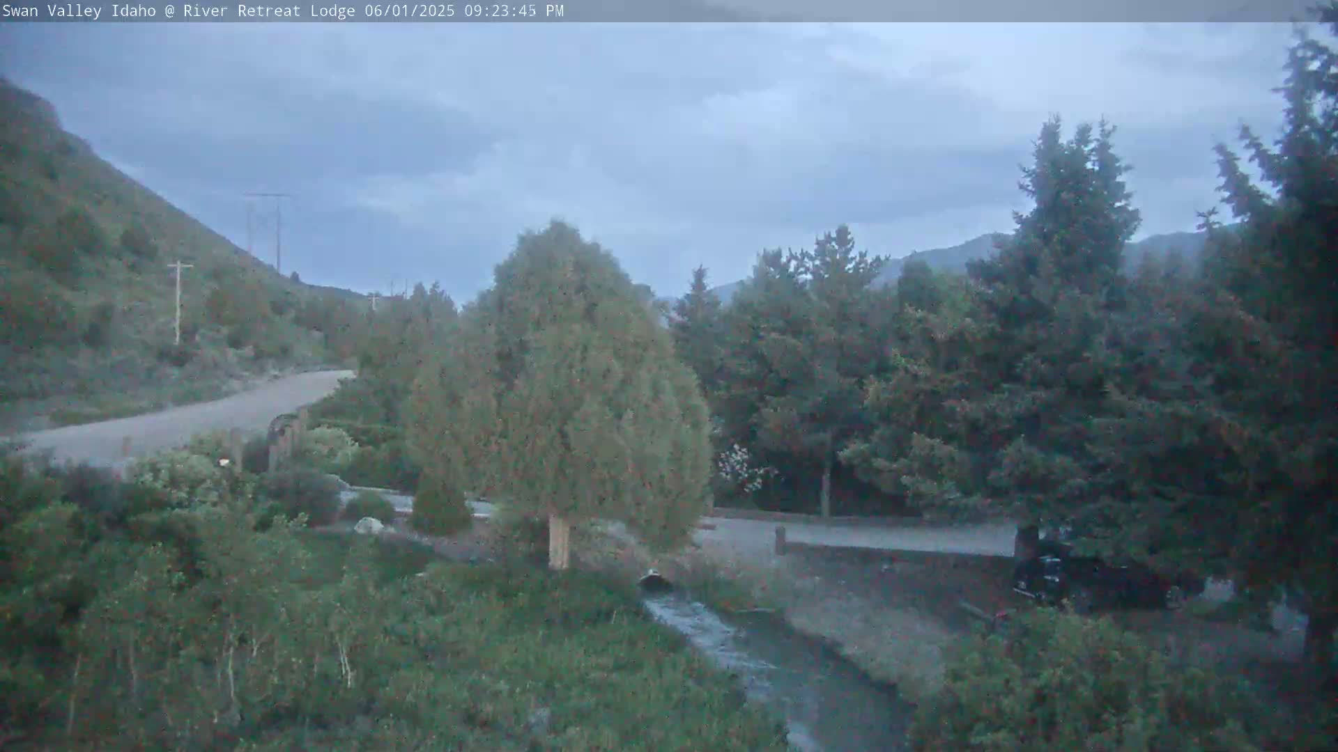 A paved road winds through a mountainous area with evergreen trees and a small stream, under an overcast sky.