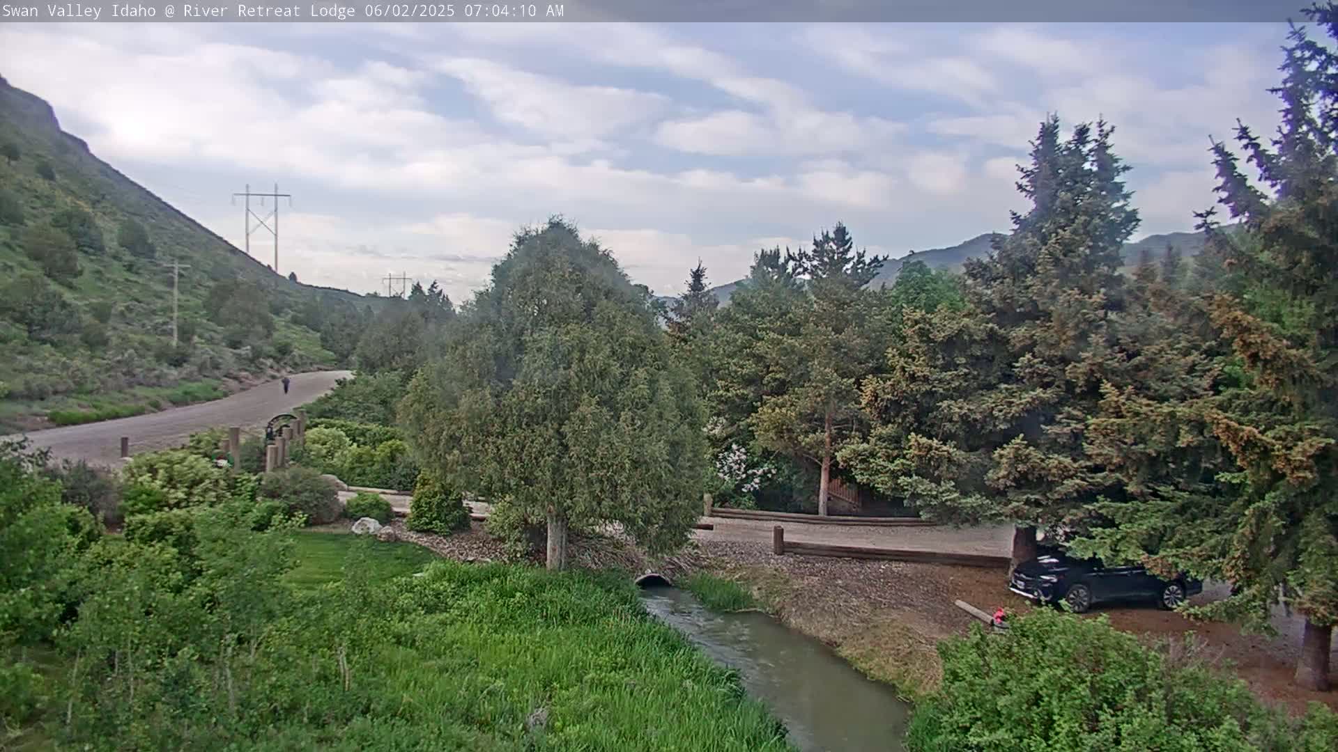 A partly cloudy day reveals a paved path winding through lush green vegetation alongside a small stream, with a car parked near tall evergreen trees and a person walking in the distance.