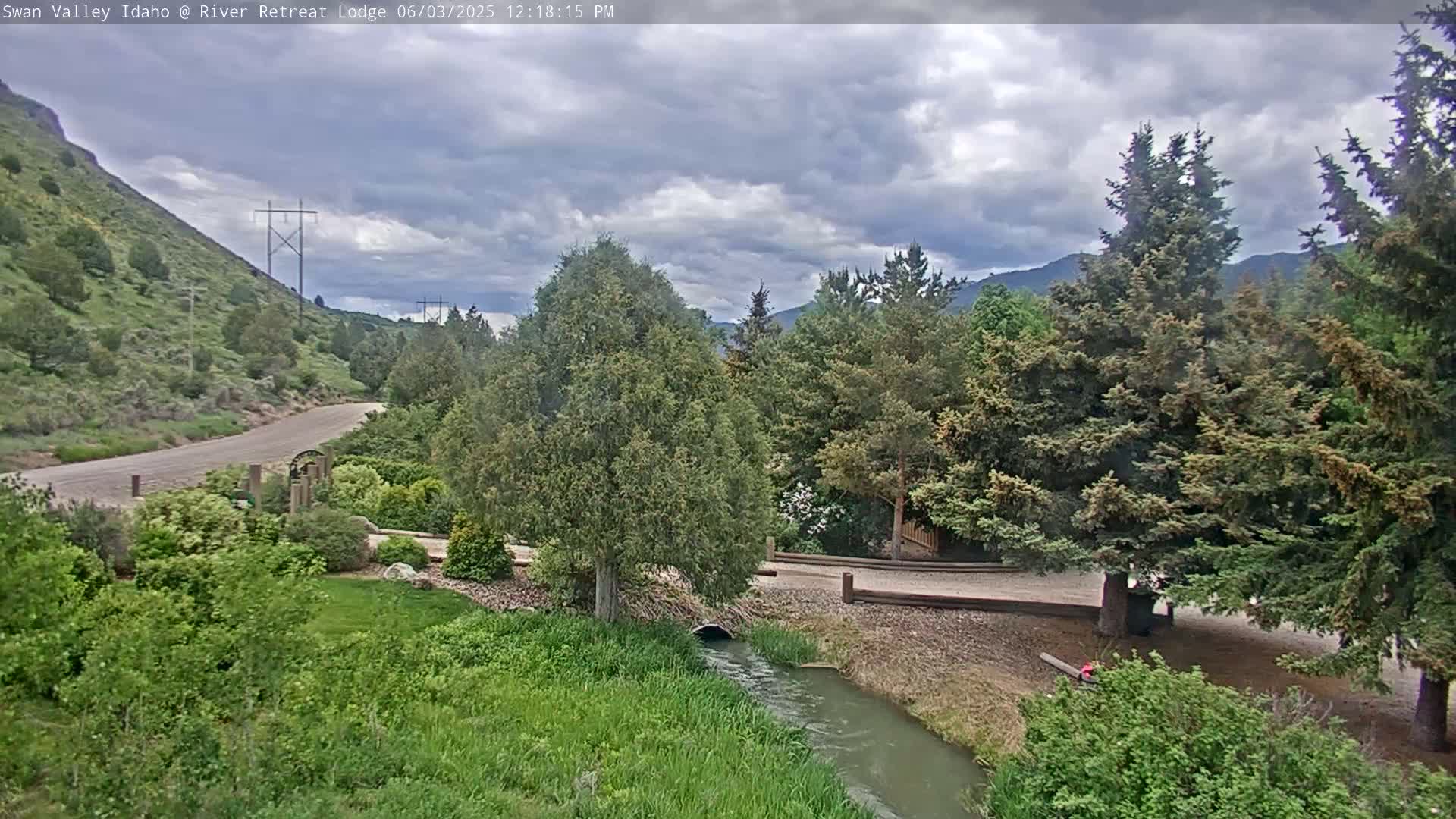 A small stream flows through a landscaped area with evergreen trees and a gravel path under a partly cloudy sky.