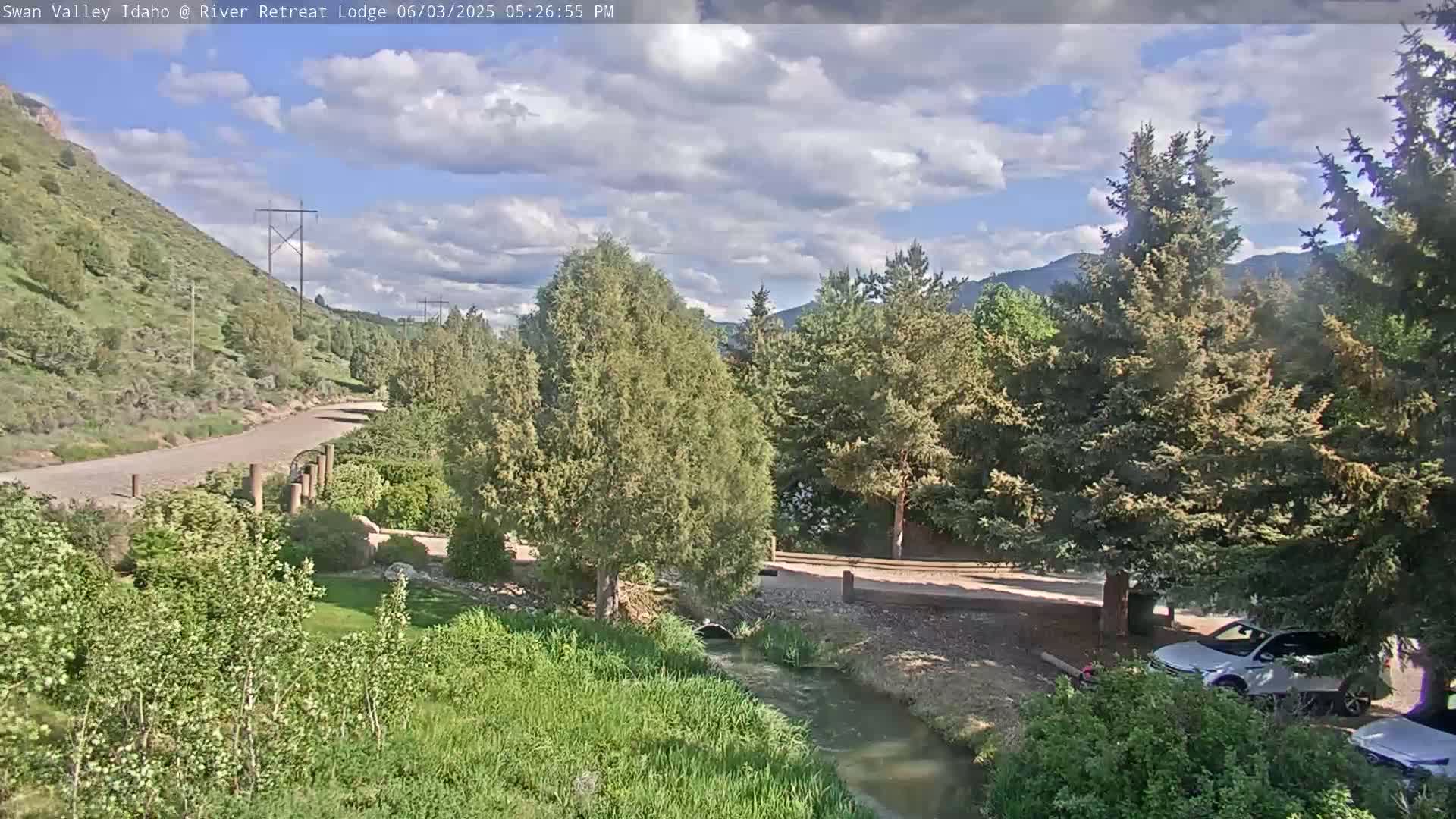 A partly cloudy afternoon scene shows a gravel road winding through a green valley alongside a small stream, with evergreen trees and mountains in the background.
