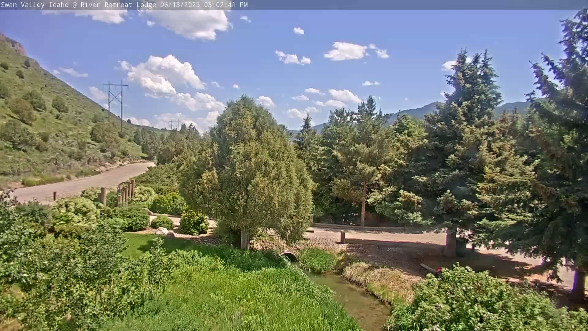 A partly sunny day reveals a paved road winding through a lush green landscape, featuring a small stream, tall evergreen trees, and a hillside in the background.