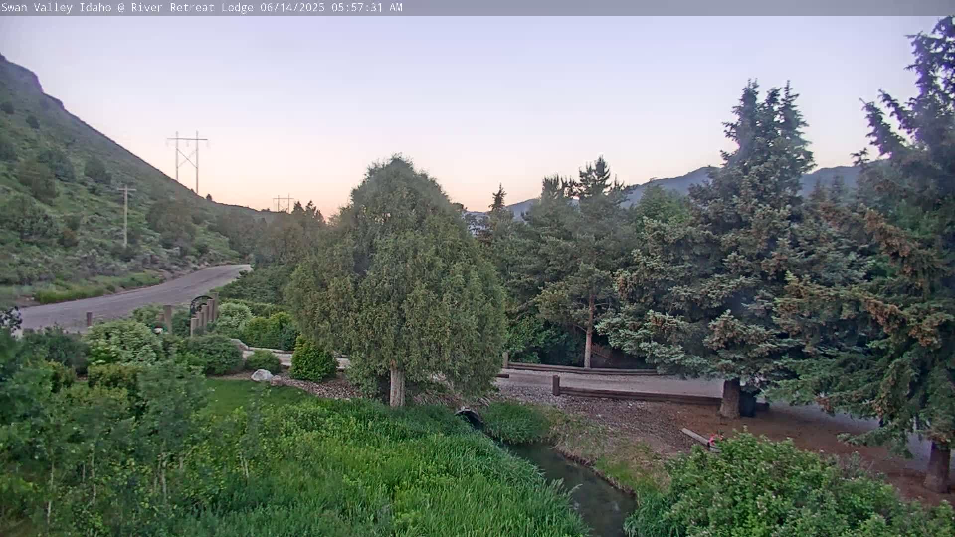 A paved road curves through a green valley at dawn, with a small stream running alongside a wooded area and a hill in the background.