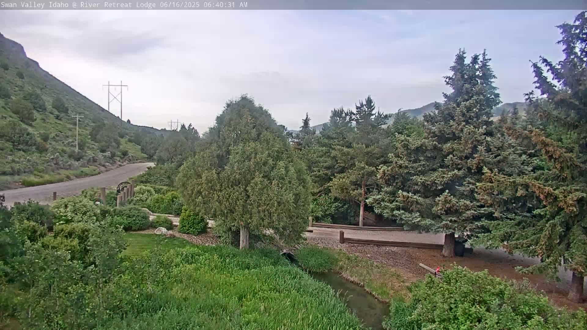 A paved road winds through a valley of green hills and evergreen trees under a cloudy sky, alongside a small stream.