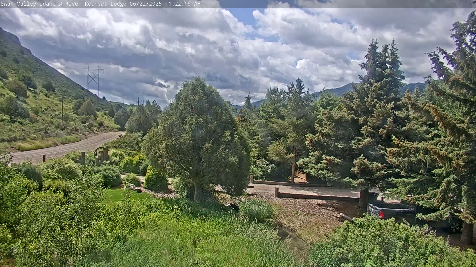 A partly cloudy day reveals a gravel road winding through a lush green valley, bordered by evergreen trees and a hillside, with a dark-colored vehicle parked near some trees.