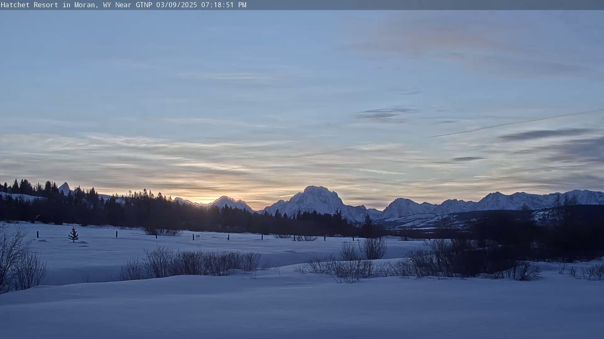 A snow-covered field stretches before a line of snow-capped mountains at sunset under a pale blue sky.