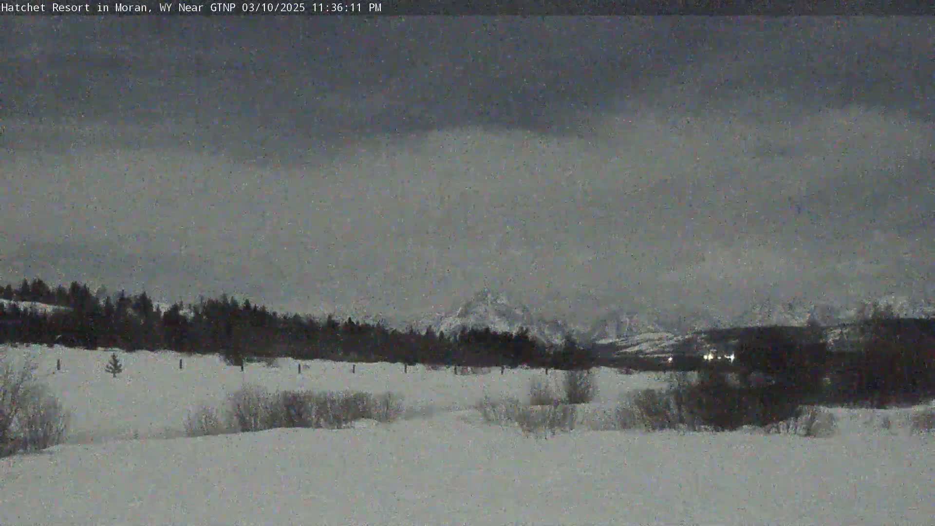 A snow-covered landscape with a dark, overcast sky and distant snow-capped mountains is visible at night.