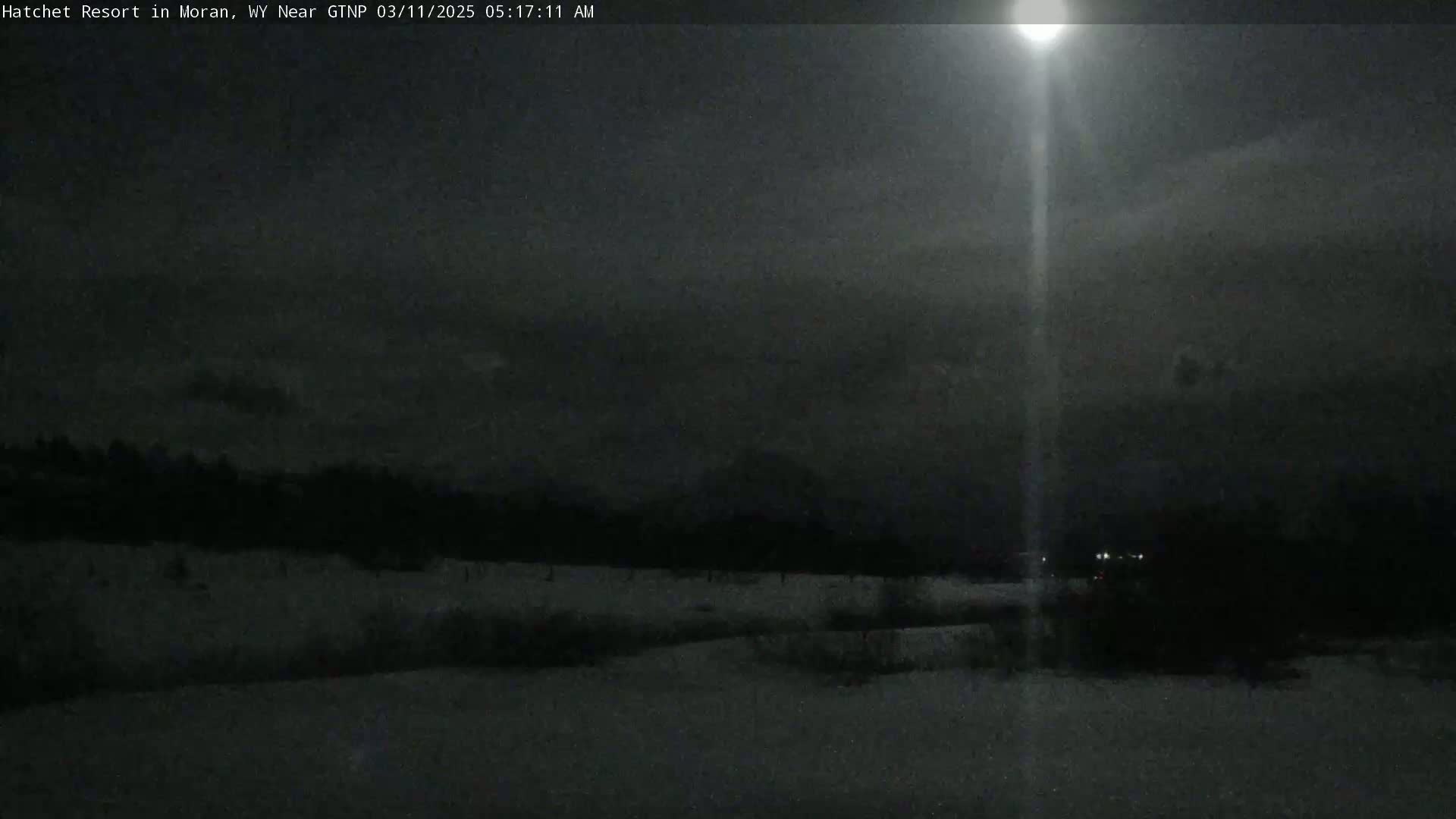 A nighttime, snowy landscape is illuminated by a bright light source, with a dark, overcast sky.